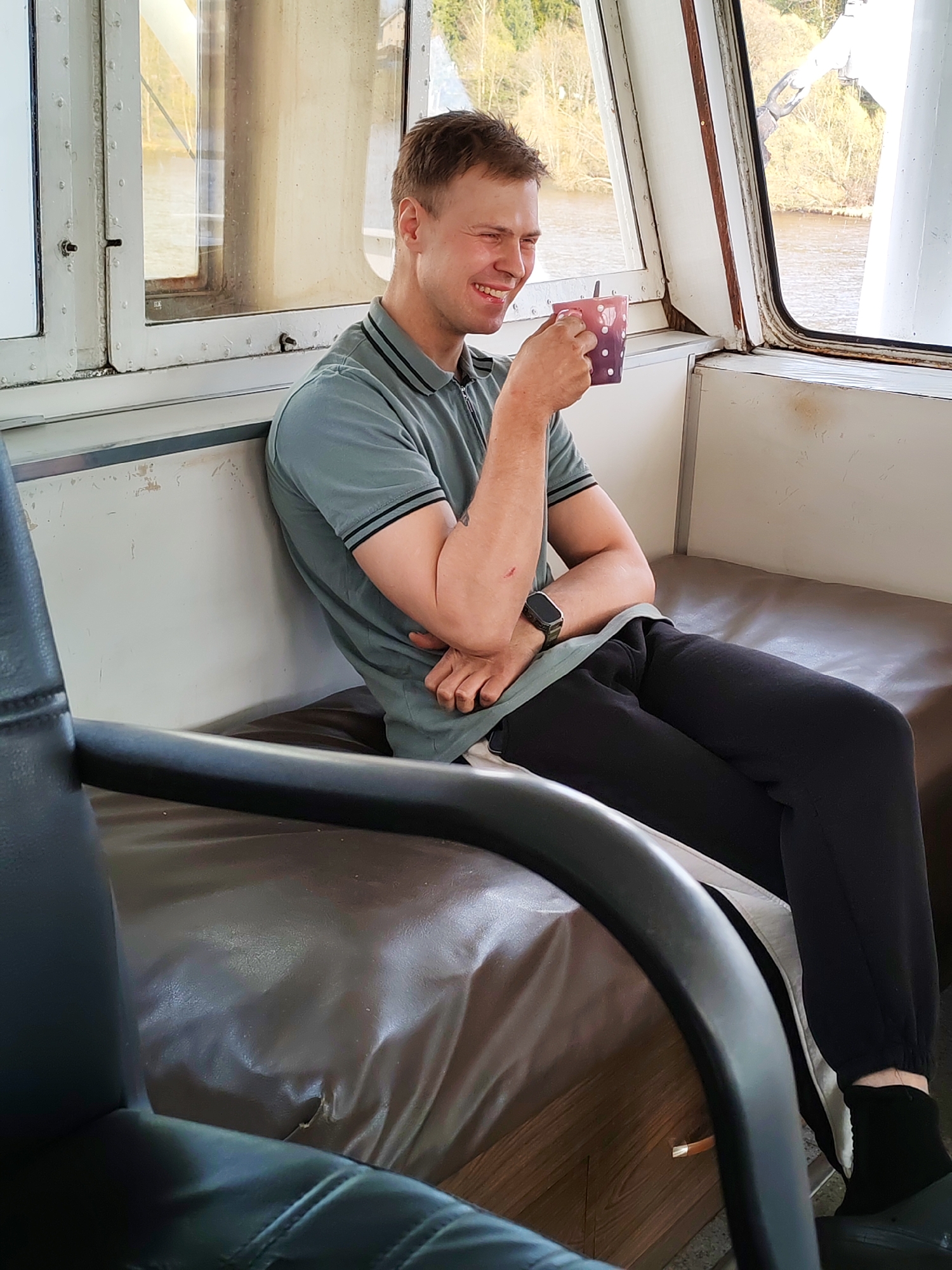 A man is seated on a dark brown bench within a cabin, possibly on a boat. He is wearing a short-sleeved gray polo shirt with a black trim, black sweatpants, and dark socks. On his left wrist, he has a watch. He is holding a polka-dotted cup, and there's a spoon sticking out of it. He is smiling slightly and looking toward the cup. The bench is located in front of a white wall and a window. The scene visible through the window appears to be a body of water with land and trees in the background. The lighting is natural. The location is identified as being in the city of Фёдоровка, Russia. A black chair is visible in the foreground.
