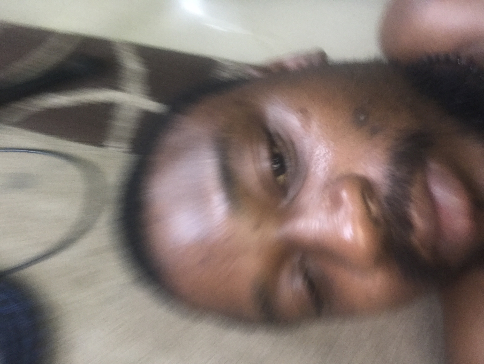 A blurry, close-up portrait features a person, likely male, with dark skin and visible facial hair around the chin and jawline. The subject's face is tilted sideways, with one eye clearly open and looking towards the camera, and a faint smile or smirk on their lips. Their skin exhibits some sheen, possibly from natural oils or lighting.
The background is highly blurred, suggesting motion blur or a shallow depth of field. Elements discernible in the background include a dark brown fabric with a lighter, geometric pattern, possibly part of a blanket or upholstery, on the upper left. A thin, dark, curved object, which could be a cable or earphone, is also faintly visible in the blurred area to the left of the person's head. The scene appears to be indoors.
The primary activity is the individual posing for a photograph. The image lacks sharp detail and is presented at an angle. There are no specific cues for time of day or weather, nor any visible text. The individual's appearance is consistent with the provided location context of Abuja, Nigeria.