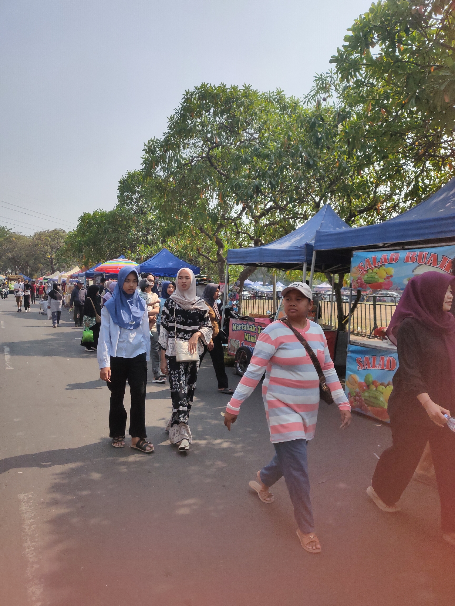 The image shows a street scene in Ngasem, Indonesia, possibly during a local market or festival. Several people are walking on a paved road lined with tents and stalls. The tents appear to be selling food and drinks, with signs visible advertising "SALAD BUAH" (fruit salad) and "Martabak Mini". The people in the foreground are dressed in casual clothing, with some wearing headscarves, suggesting a predominantly Muslim population. The weather appears to be sunny and warm, with clear skies and trees lining the street. The overall atmosphere seems lively and bustling, indicative of a community gathering.