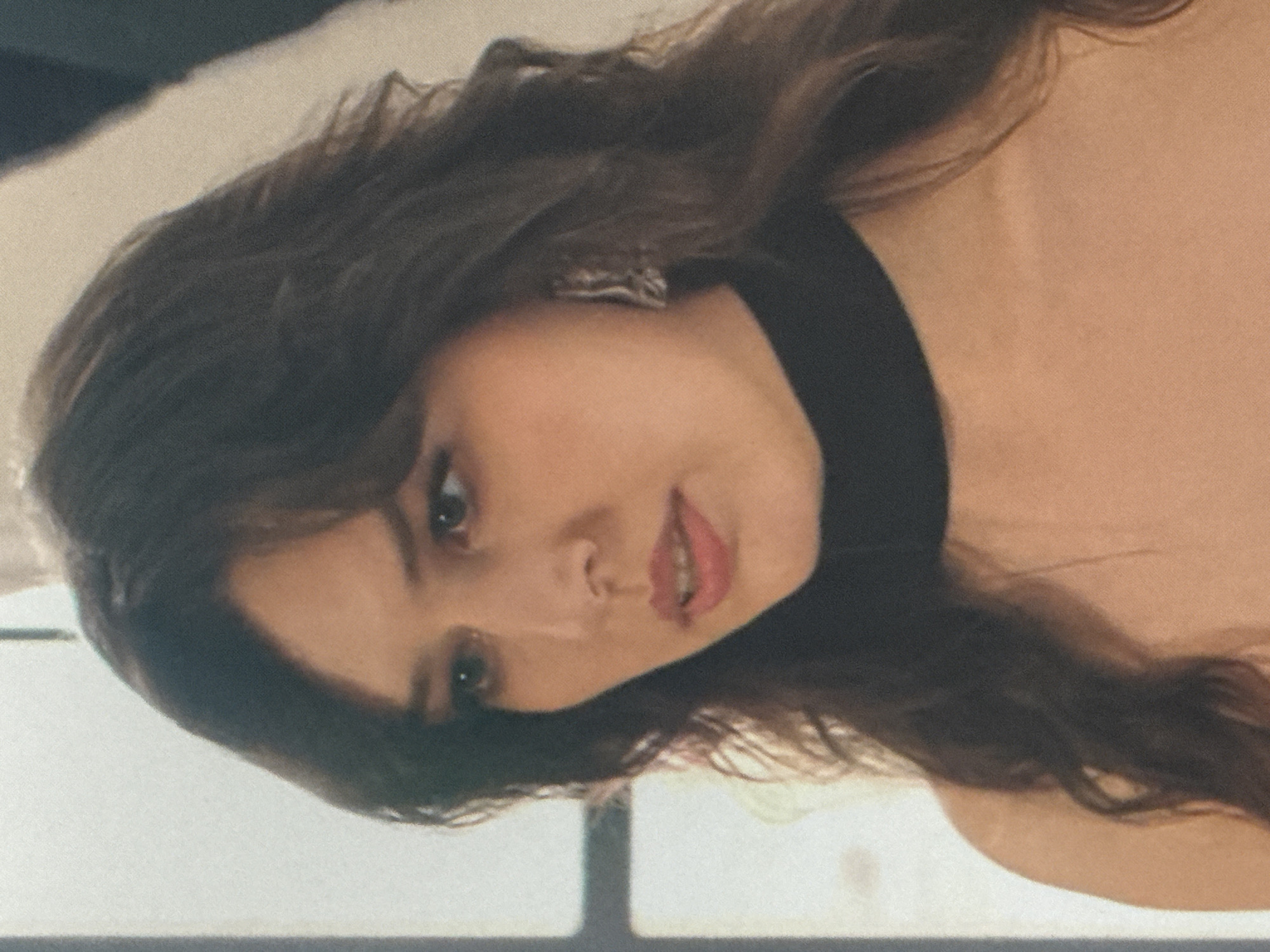 A medium shot captures a woman from the chest up, with her head slightly turned and looking downward. She has long, dark, wavy hair that drapes over her shoulders. Her face is framed by her hair, and she wears a black choker necklace. She has large, dark eyes and is wearing a noticeable silver earring on her right ear. The background is out of focus, showing a soft, neutral tone with a hint of a window or light source on the left side, and a darker shape on the right. There are no identifiable landmarks or clues to suggest the location is in Saratov, Russia.