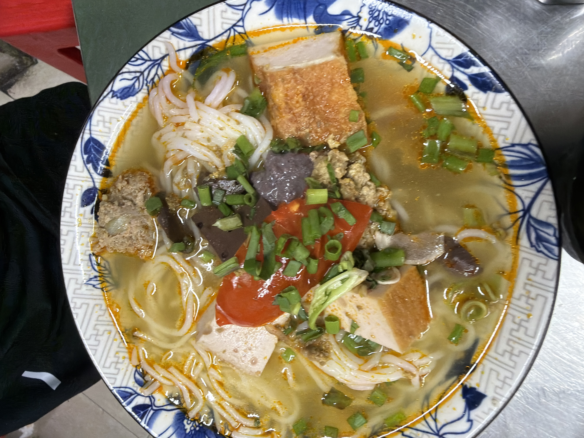 A close-up, overhead shot captures a vibrant bowl of Vietnamese noodle soup, likely *Bún riêu* or *Bún mọc*, served in a white ceramic bowl with a distinctive blue and white patterned rim featuring both floral and geometric designs. The golden-hued broth contains white rice vermicelli noodles and a variety of toppings. Visible ingredients include a large piece of crispy pork belly with crackling skin, crumbled ground pork or a pork patty, dark cubes of blood pudding, slices of white pork loaf (*chả lụa*), bright red chopped tomatoes, and a liberal garnish of chopped green onions and fresh herbs. The bowl rests on a surface that is partially metallic, resembling a kitchen counter or table, and partly a dark green material. A glimpse of dark fabric is visible on the bottom left. The dish is presented as a finished meal, typical of Vietnamese cuisine, consistent with the location context of Đà Nẵng, Vietnam. No people or visible text are present.