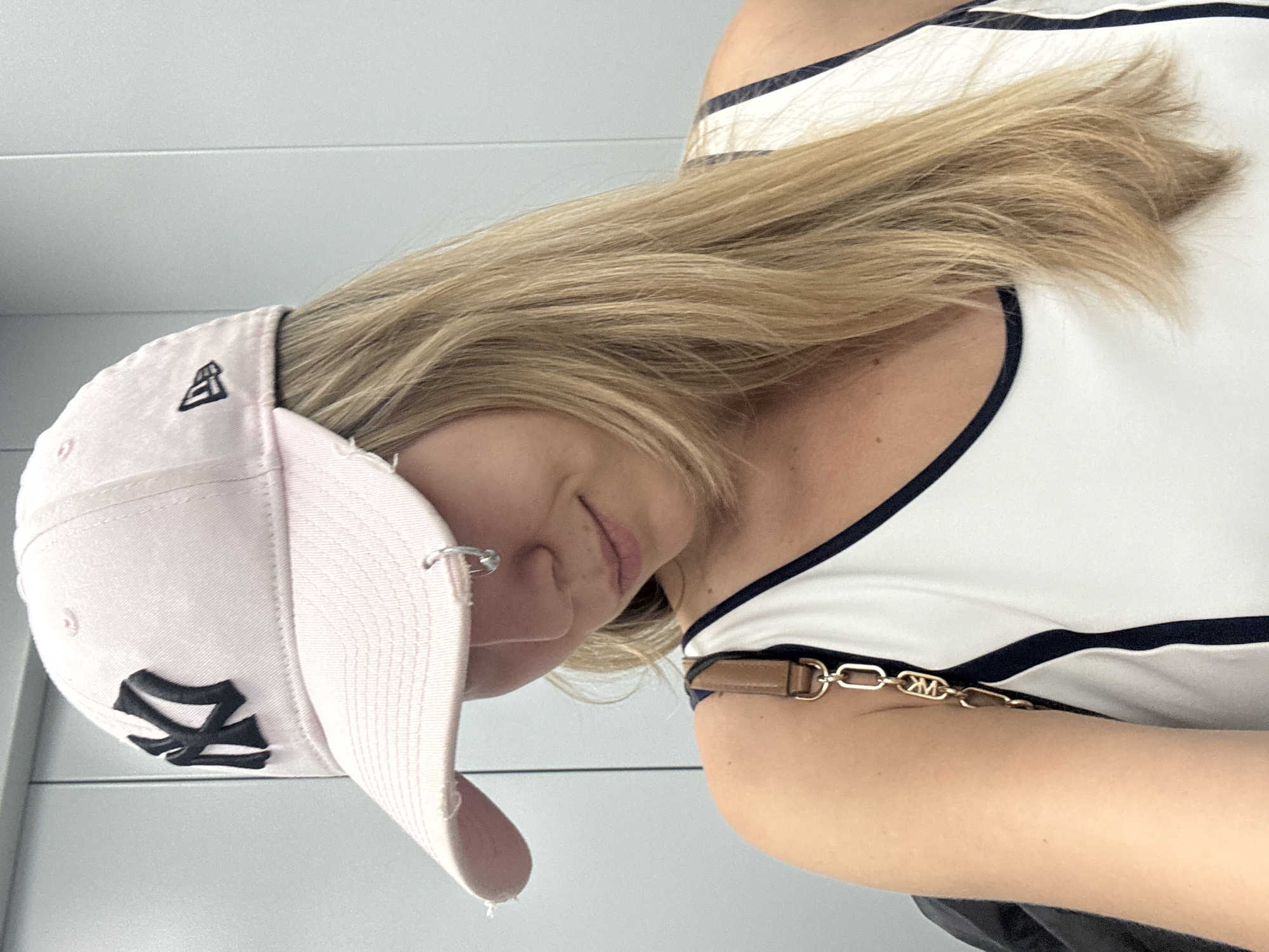 A young Caucasian woman, with blonde hair and fair skin, is shown from the chest up in a selfie, looking down and to the right. She is wearing a light pink baseball cap with a black "NY" logo and a New Era logo. She has a silver hoop earring in her left ear. She is also wearing a white tank top with navy blue trim. A brown leather purse strap with a gold chain and a "MK" logo is visible over her left shoulder. The background appears to be a light grey wall, possibly in a hallway or elevator. The lighting is bright and even. The woman's expression is relaxed and slightly smiling.