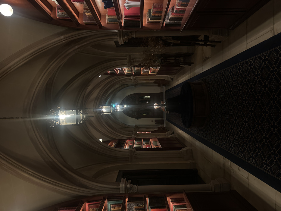 The scene depicts a long, dimly lit, gothic-style library hallway in Cernay-la-Ville, France. A vaulted stone ceiling spans the corridor, supported by stone pilasters. Light-toned stone tiles cover the floor, centrally traversed by a dark blue runner rug featuring a repetitive geometric pattern. Built-in wooden bookshelves, illuminated by integrated warm strip lighting, line both walls, displaying numerous antique-bound books of various sizes and colors. Multiple ornate, dark metal lanterns with glass panels hang sequentially from the ceiling, providing primary illumination. Approximately midway down the visible hallway, a circular wooden ottoman with a dark cushion is centrally positioned on the rug, supporting a candelabra with three lit candles. At the far end, a substantial, dark, carved wooden door is visible. No persons or actions are present within the scene.