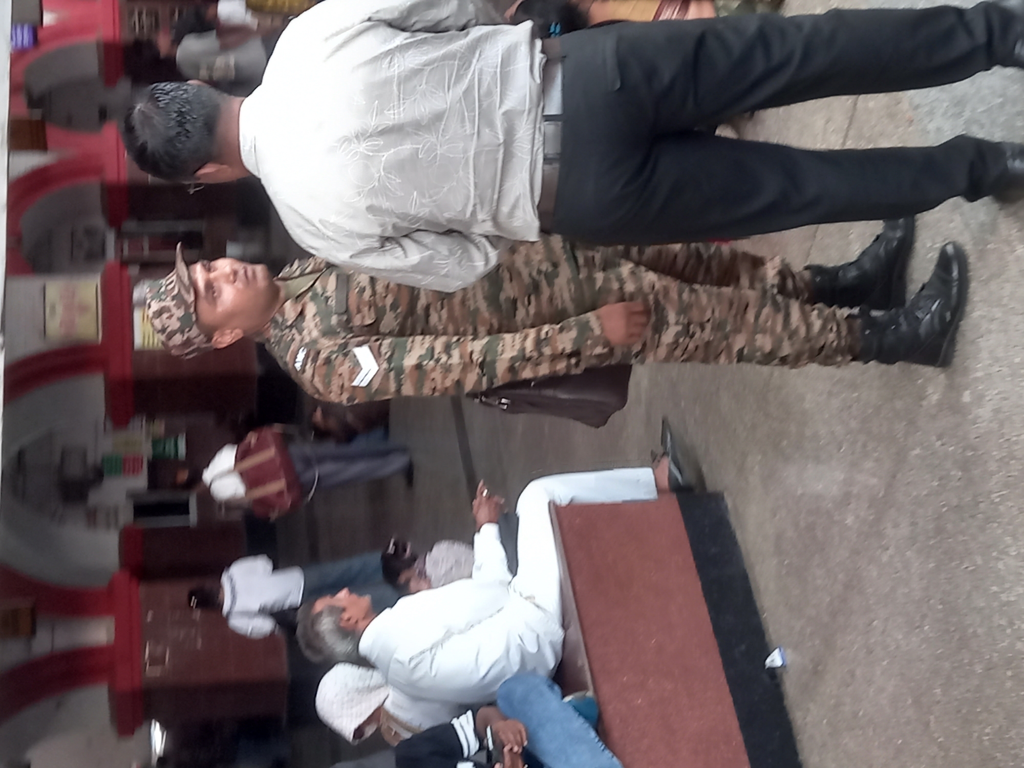 The image depicts an indoor scene in what appears to be a public space in Jhansi, India. In the foreground, an older man is lying on a cot with a white sheet, wearing a white kurta and blue jeans. Next to him sits another individual. Standing nearby are two more figures: one is in a camouflage uniform and cap, wearing black boots, and the other is in a white shirt and black pants. The camouflage-clad figure has a shoulder bag. The setting includes dark wooden storage cabinets and red arched doorways. Other people are visible in the background. The location is possibly a government office or a public waiting area.