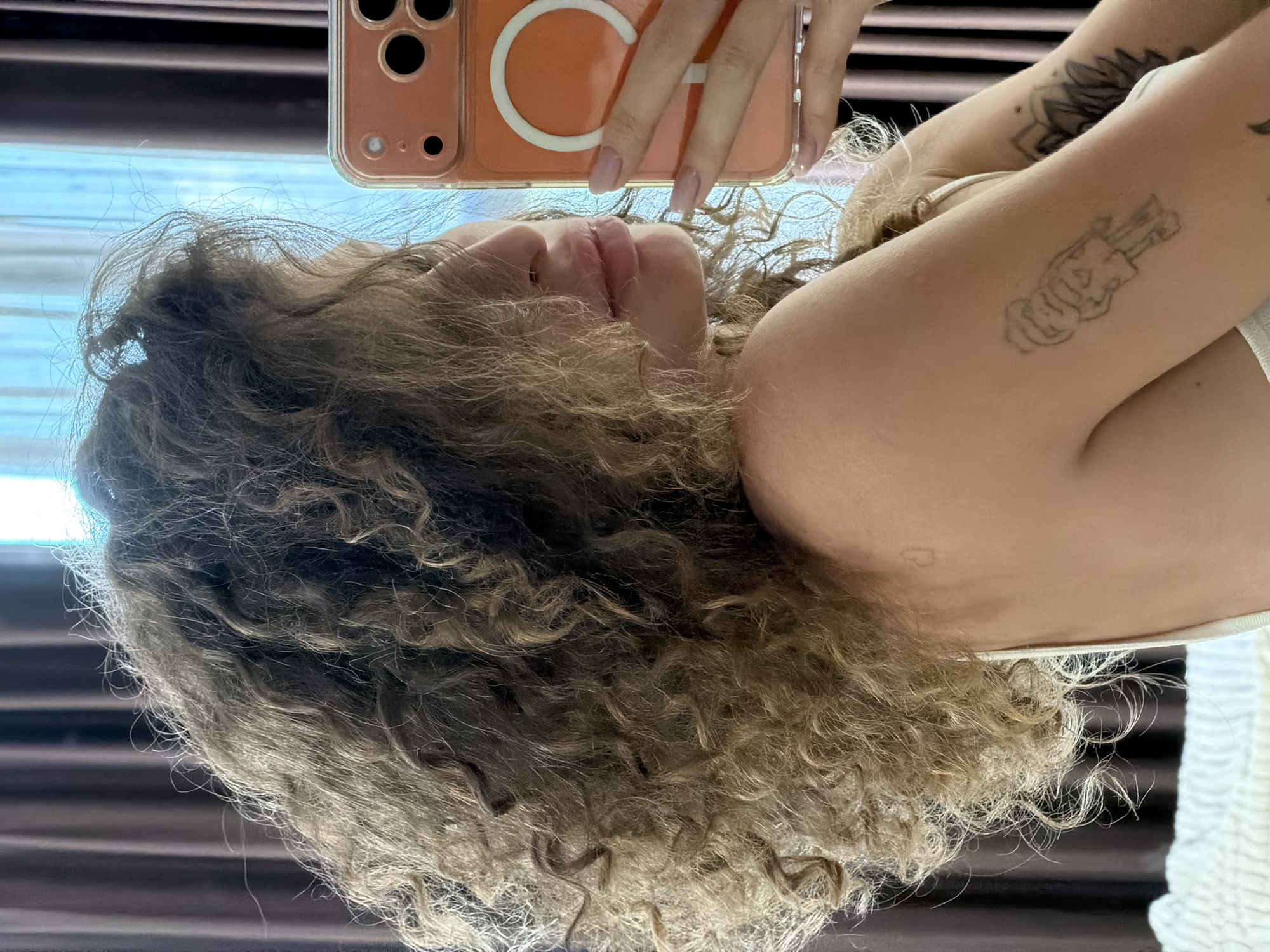 A woman with curly blonde hair is taking a selfie with an orange iPhone. The woman is wearing a white tank top. The background includes brown curtains and a blue window. Tattoos are visible on her arm. The location in North Kuta, Indonesia is not verifiable from the image.