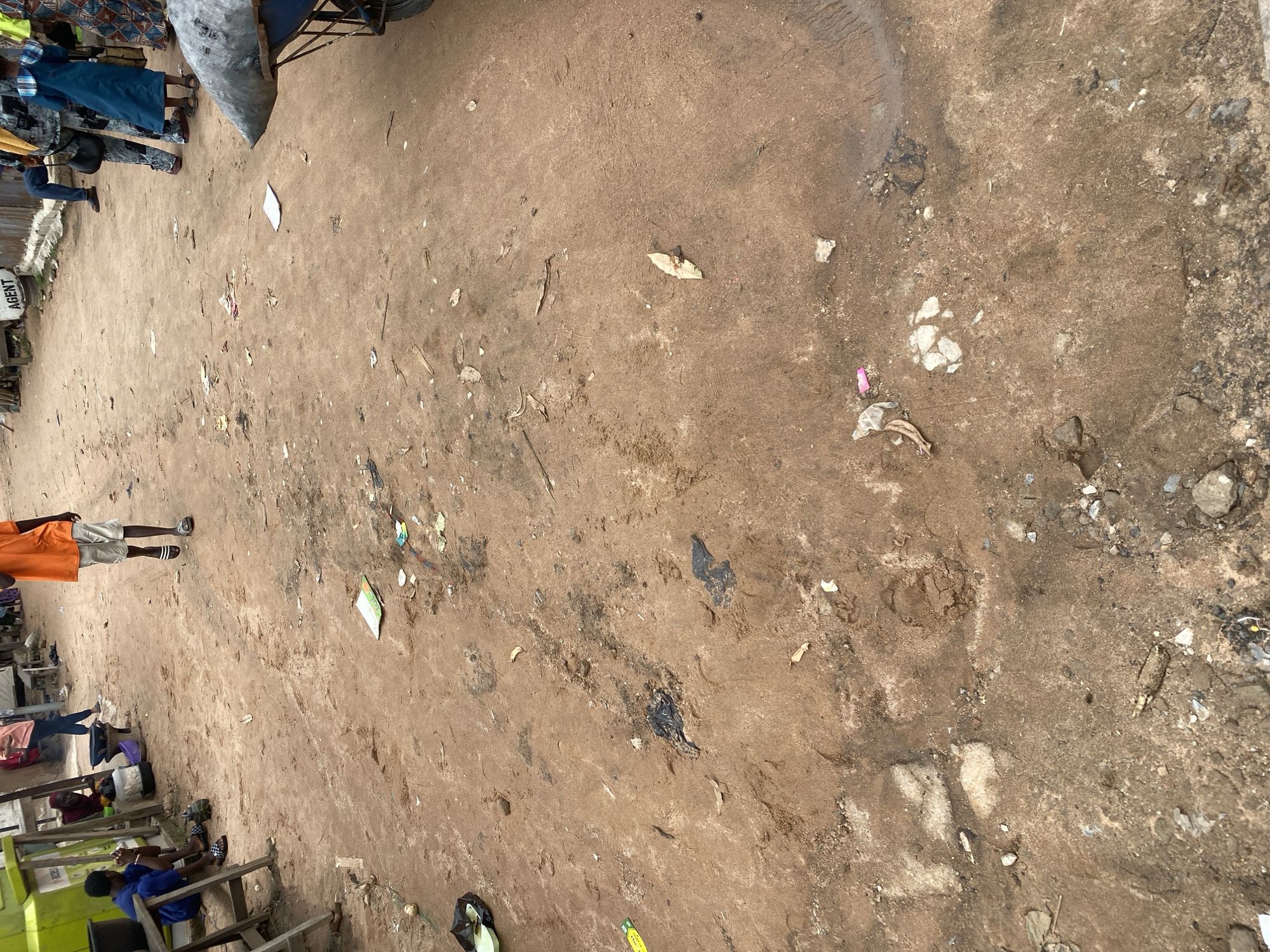 This media file captures an outdoor scene in Jalingo, Nigeria, likely a market or busy thoroughfare. The ground is primarily dirt, with patches of mud and scattered debris, including plastic bags, paper scraps, and stones, suggesting a somewhat unkempt environment.
Several people are visible. In the background, a group stands near a cart with a large, grey, conical object on it, and a black tire is visible. One person in the foreground is wearing an orange shirt and light-colored shorts, standing with their legs apart, and appears to be looking down. To the left, a woman in a blue dress and a yellow scarf is also visible, as well as other individuals in the distance. In the lower left of the frame, a few people are seated on wooden structures, possibly stalls or benches. One person is wearing a blue shirt.
The scene suggests a daytime setting, with ambient lighting that doesn't indicate harsh shadows. The overall impression is one of everyday activity in a public space. A white sign with black text that reads "AGENT" is visible on the left side of the frame.
