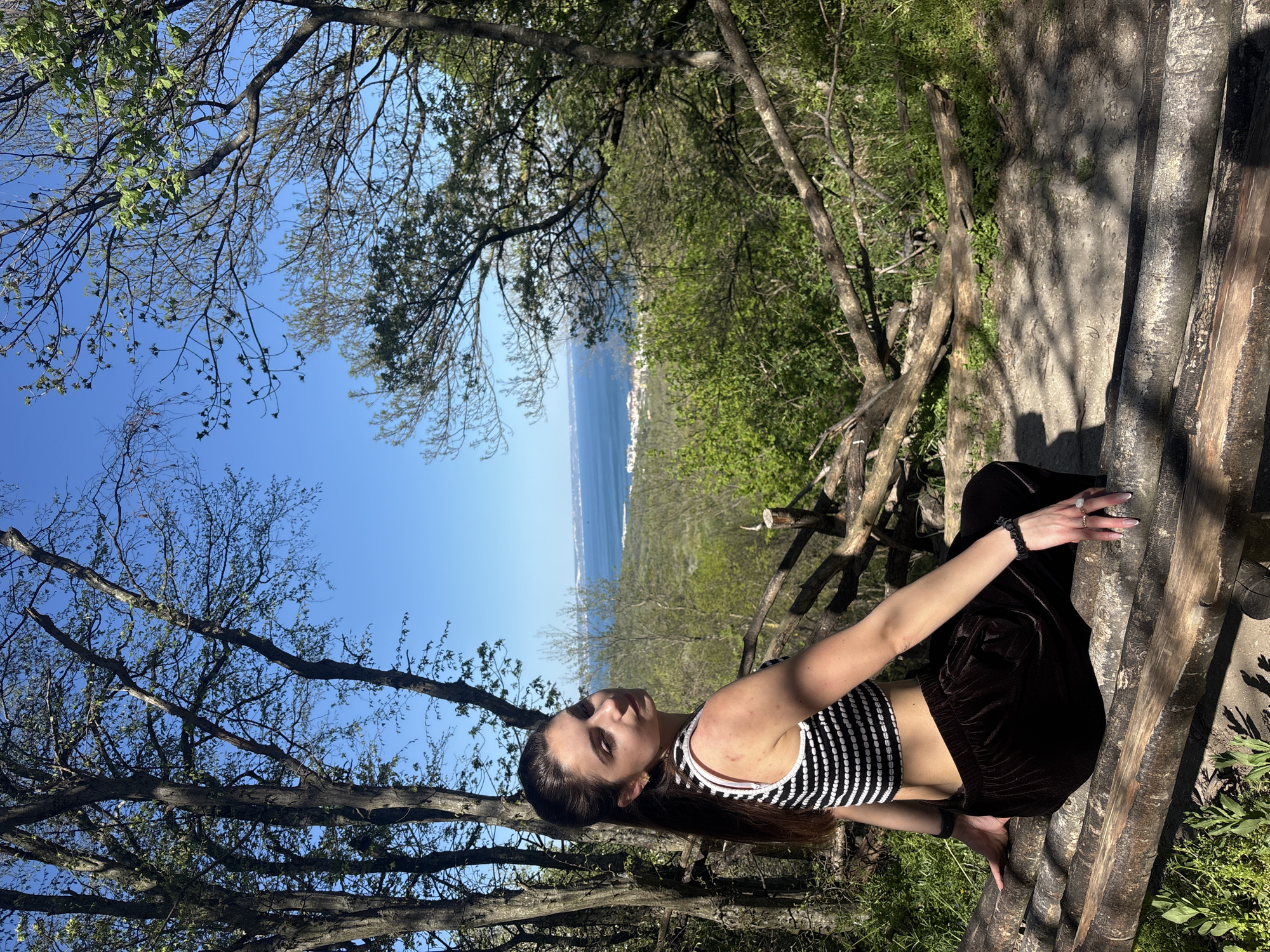 A woman is perched on a wooden structure, seemingly a railing or a bench, in a natural setting overlooking the coastline. The woman is wearing a black and white striped tank top and brown pants. She is facing the camera with a slight tilt of her head. Surrounding the woman are trees with a view of the sea in the background. The location is likely near Varna, Bulgaria, due to the presence of a coastline with distant urban features suggesting a coastal region. The image seems to be taken on a bright day with clear skies.
