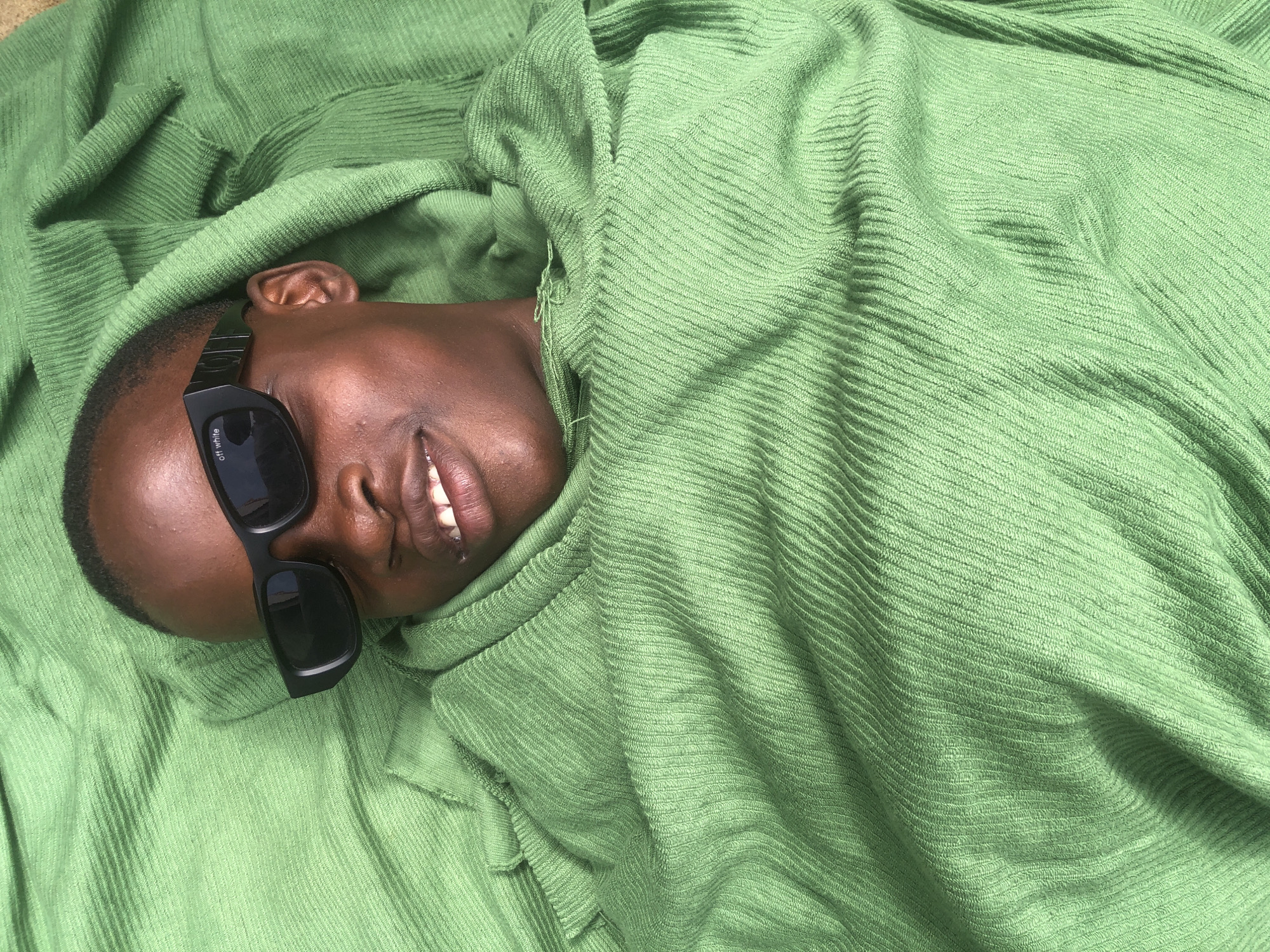 In Ado, Nigeria, a person with dark skin and short hair wears black sunglasses and is wrapped in a green, ribbed fabric. The fabric is draped around their head and shoulders, with visible folds and texture. The person's mouth is slightly open, revealing teeth, and they are looking upwards. The lighting suggests an outdoor environment, possibly during the day.