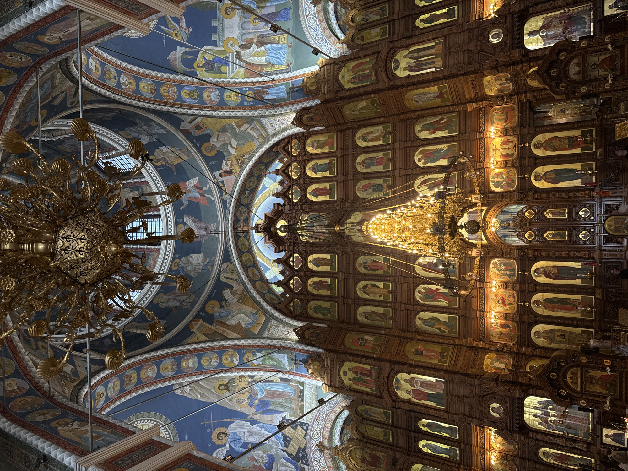 The image depicts the interior of a Russian Orthodox church, possibly in Nizhny Novgorod, Russia. The scene is focused on the ceiling, which is divided into two distinct sections. The left section features an ornate, golden chandelier hanging in front of a blue-painted dome adorned with religious figures and circular medallions. The right section showcases a wooden iconostasis panel covered in framed religious icons, with another chandelier hanging in front. The overall impression is one of grandeur and religious significance.