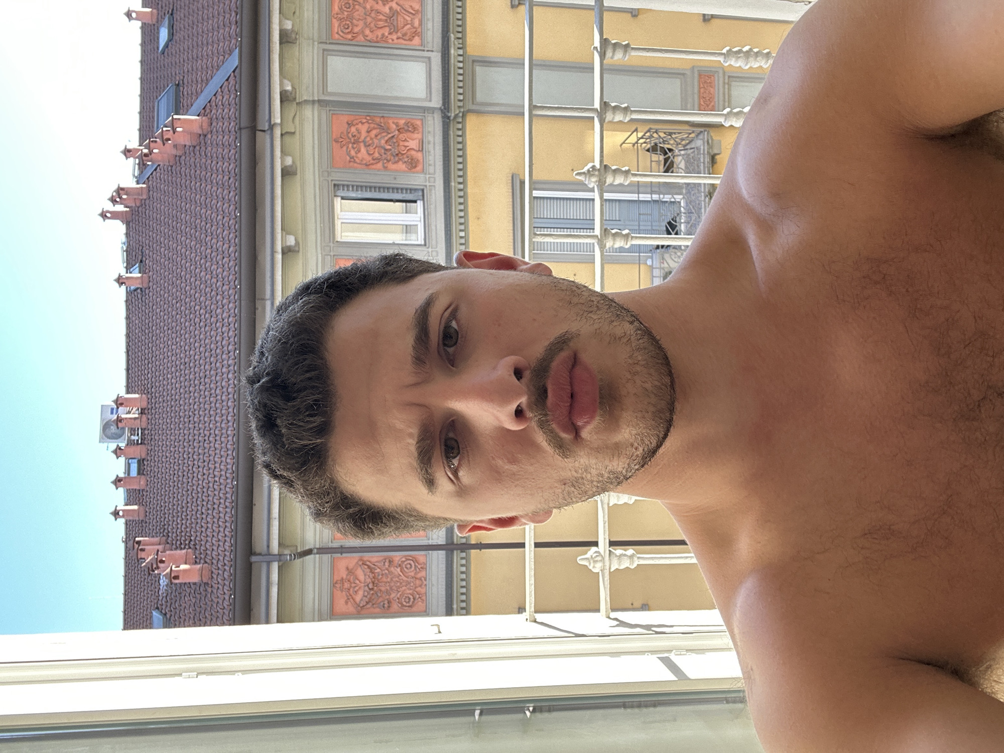 A medium-shot, selfie photograph depicts a shirtless, light-skinned male posing in front of a window with a metal railing and architectural buildings in the background. The buildings are located in Milan, Italy, as evidenced by the chimney stacks. The man has short, dark hair, light stubble, and his lips are pursed. His right shoulder and chest are visible. The foreground includes the bottom of the window. The buildings in the background are multi-story and exhibit ornate facades with windows and other architectural details. The sky is light blue.