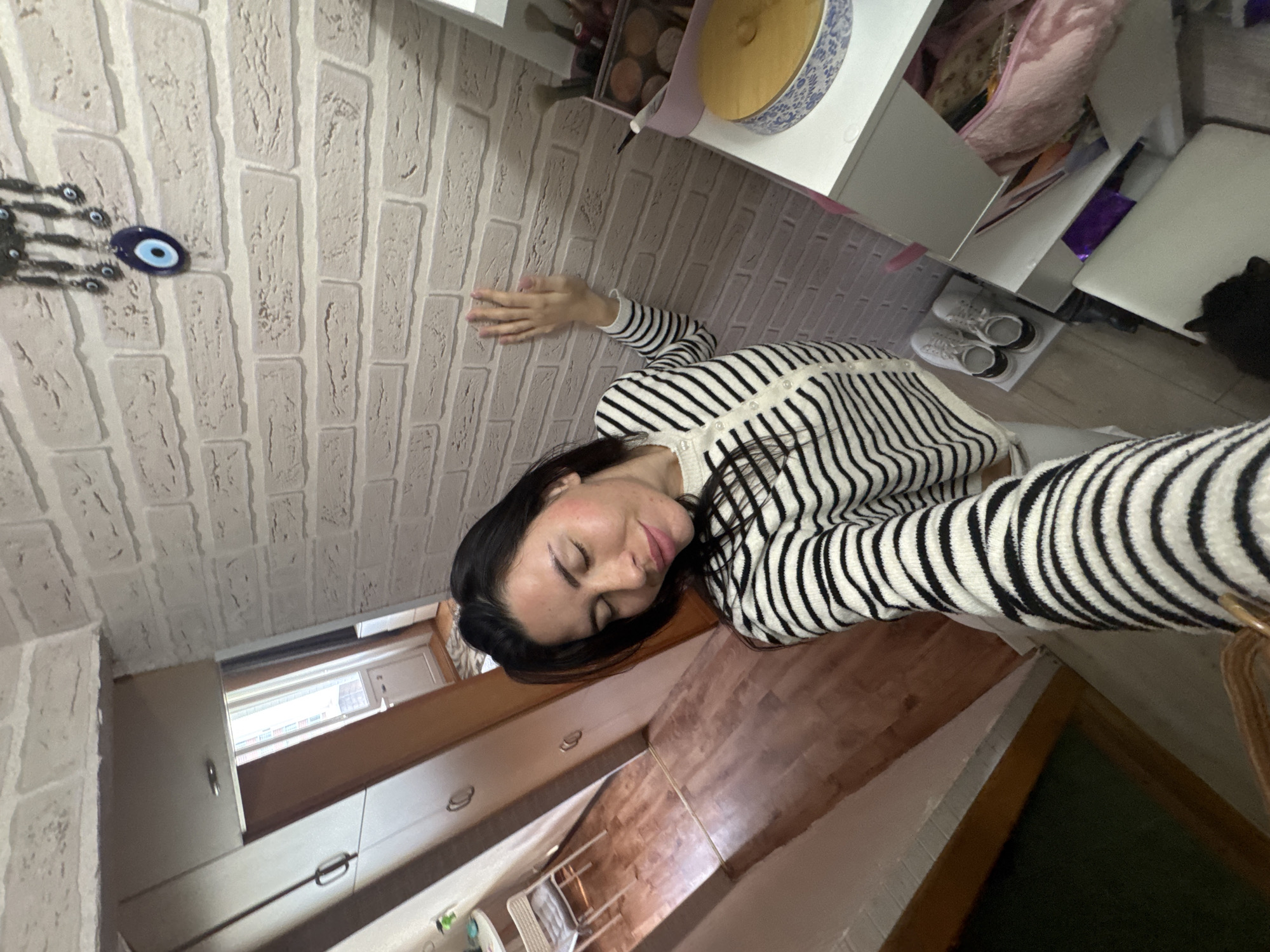 A woman with dark hair, wearing a black and white striped cardigan, is taking a selfie indoors. She has her eyes closed and her head tilted. Her left hand is resting on a brick-patterned wall. In the background, there is a hallway with wooden flooring, a tall wardrobe, and a doorway leading to another room. To the right of the woman is a white shelving unit with various items, including makeup and a decorative blue and white patterned container. A pair of white sneakers is placed on the floor next to the shelving unit. A black cat is visible at the bottom right of the image. The overall environment appears to be a residential interior.