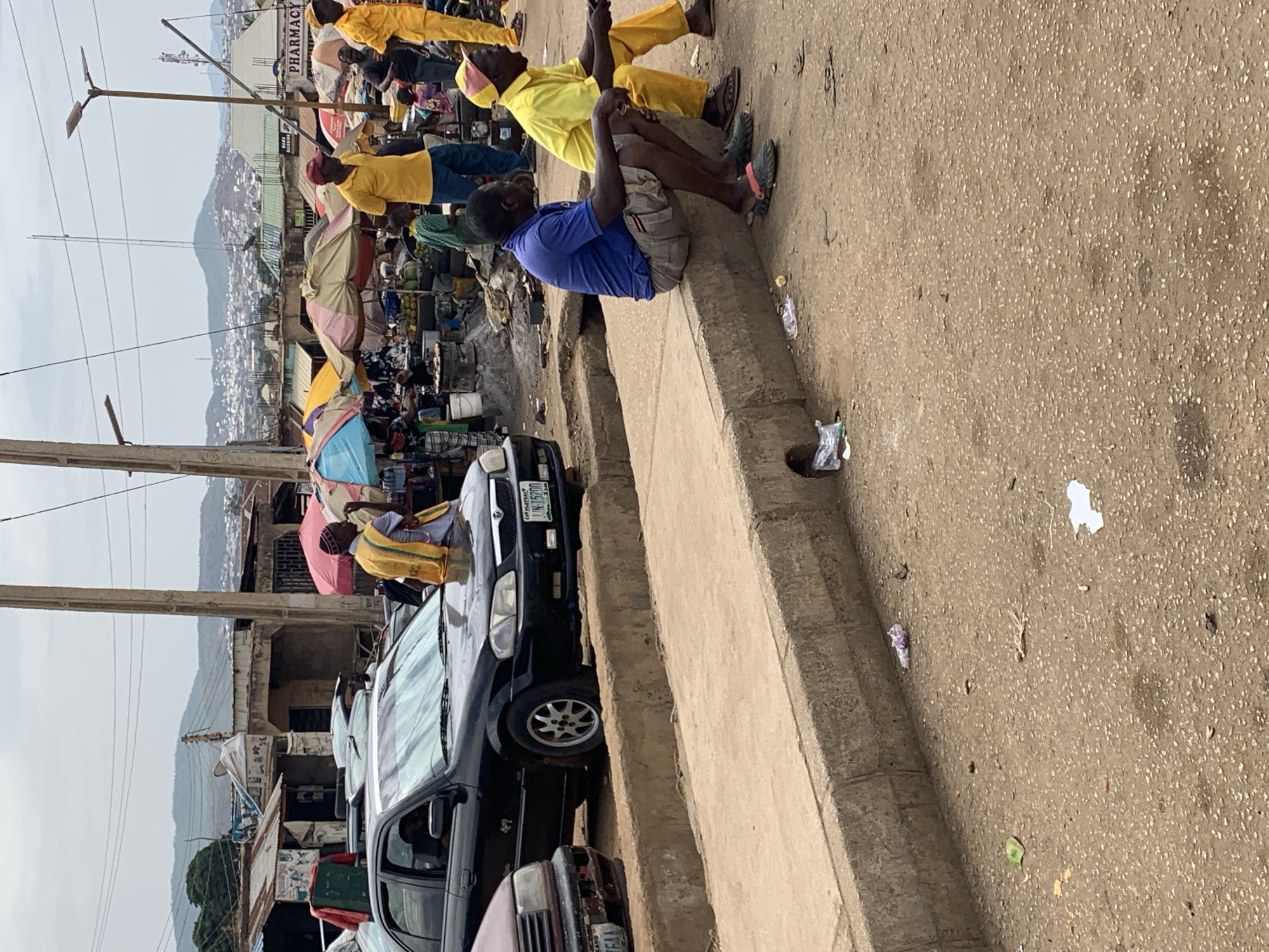 **Location:** Rafin Par, Nigeria.
**Scene:** The image captures a street scene in Rafin Par, Nigeria. Along the roadside, a group of people are visible. Several individuals are sitting on a raised concrete curb or ledge, while others stand nearby or are partially obscured by umbrellas and makeshift shelters.
**People:**
* Several people are seated along the concrete curb. One is wearing a blue shirt. Several others are wearing yellow.
* There are additional people standing in the background, mostly obscured by vendor stalls and umbrellas.
**Objects:**
* Cars: A line of parked cars occupies a significant portion of the street. License plates are visible.
* Vendor Stalls: Numerous vendor stalls or makeshift shops line the street, covered by colorful umbrellas.
* Power lines are running along the left side of the image.
* Mountains are visible in the background.
**Environment:**
* The street is paved.
* The weather appears overcast or hazy.