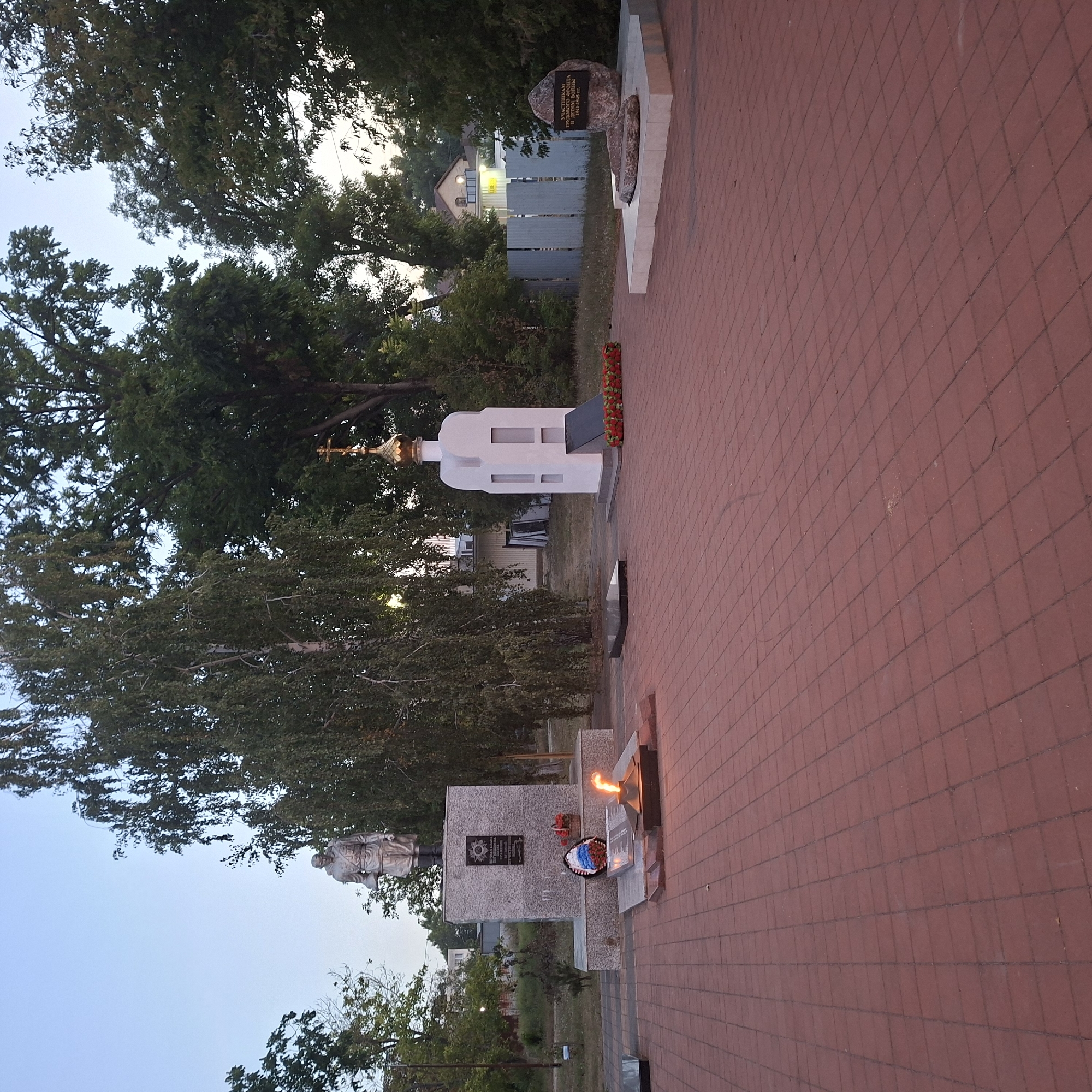 The image captures a solemn memorial site in Anapskaya, Russia, likely dedicated to fallen soldiers. In the foreground, a large, red brick paved area stretches out, leading the eye towards several commemorative elements. To the left, a statue of a soldier stands stoically atop a granite monument inscribed with text. In front of this monument, a wreath of red, white, and blue flowers lies beside an eternal flame, its orange glow a stark contrast to the twilight sky. Further to the right, a white, chapel-like structure with a golden dome and a cross stands, adorned with a red wreath at its base. Beyond these main features, trees with dense foliage frame the scene, and a subtle hint of buildings and a grey fence can be seen in the background. The overall atmosphere is one of remembrance and quiet reflection.