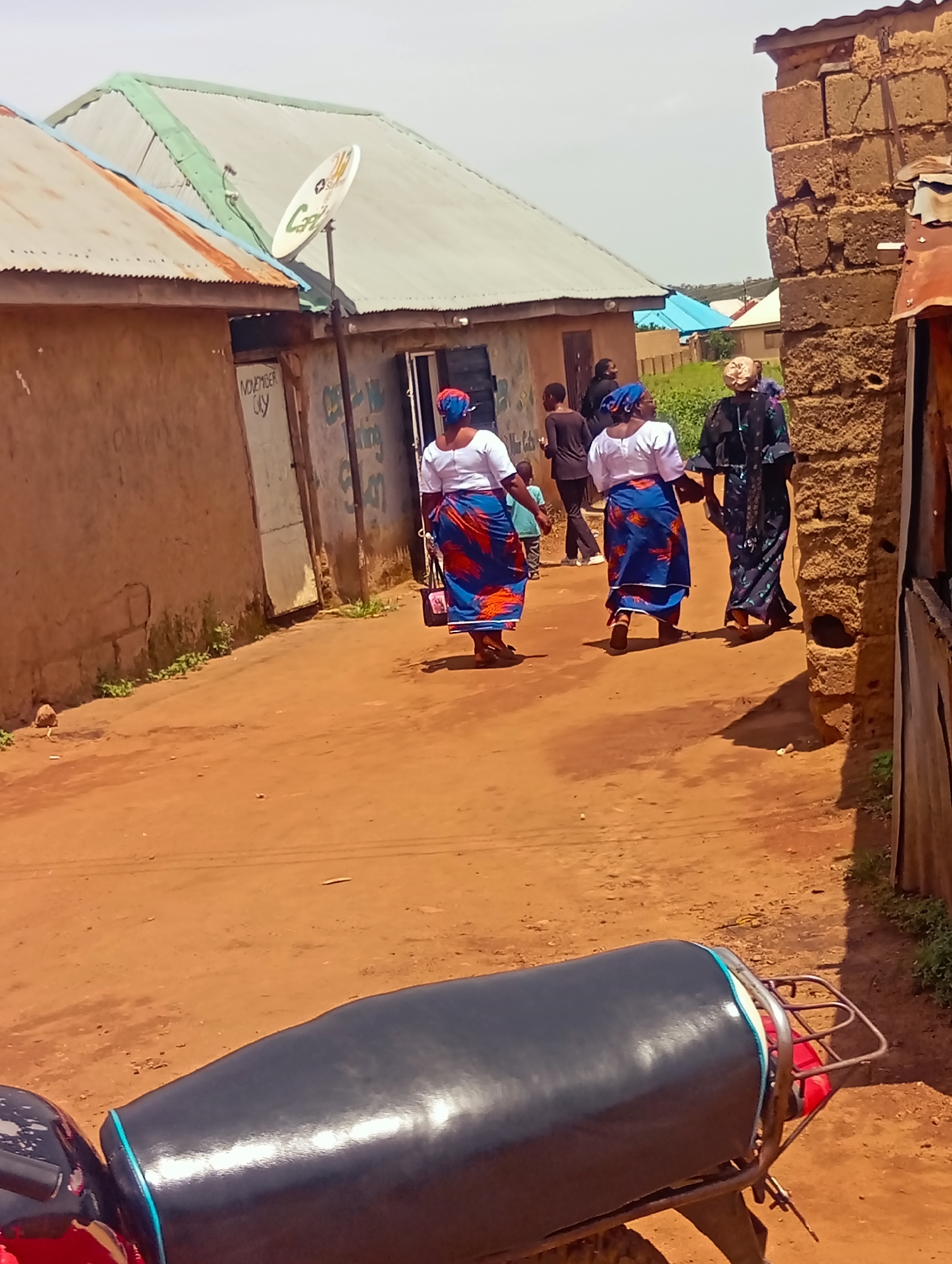 The image captures a dirt path in Du, Nigeria, lined with rustic buildings. In the foreground, the rear section of a motorcycle is visible. Further down the path, three women are walking away from the camera. The woman on the left wears a white top and a vibrant blue and red patterned skirt. The middle woman is also in a white top and a similar blue and red patterned skirt, with a blue headscarf. The woman on the right is dressed in a dark patterned robe and a light-colored headscarf. Behind them, other individuals can be seen, including a child. The buildings have corrugated metal roofs, some of which are green, and the walls are made of mud or stucco. A satellite dish is mounted on one of the roofs. The ground is a reddish-brown dirt, and sparse vegetation grows along the sides of the path. The sky is a pale blue, indicating an outdoor, daytime scene.