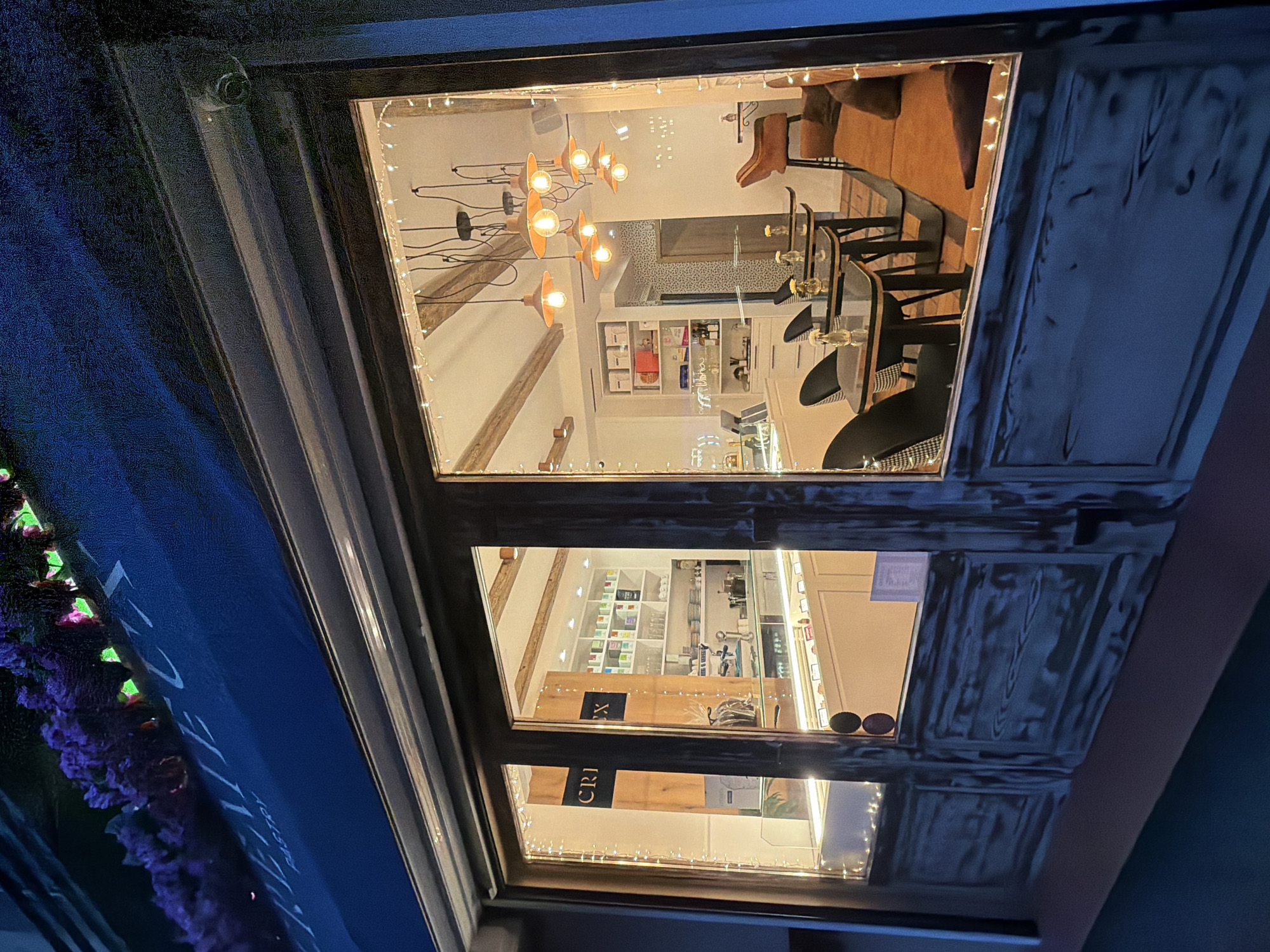 A multi-tiered window display, photographed at night, reveals the interior of a shop. The upper level of the display features a light fixture with multiple orange bulbs, seating including a brown leather chair and several black chairs at glass tables, and a white shelving unit against a patterned wall. The middle level showcases various products on display shelves. The lower level contains a sign with the word "CRIX" visible. Fairy lights are strung around the edges of the window frames. A dark blue awning with text is partially visible on the left side of the frame. The location is Sofia, Bulgaria.
