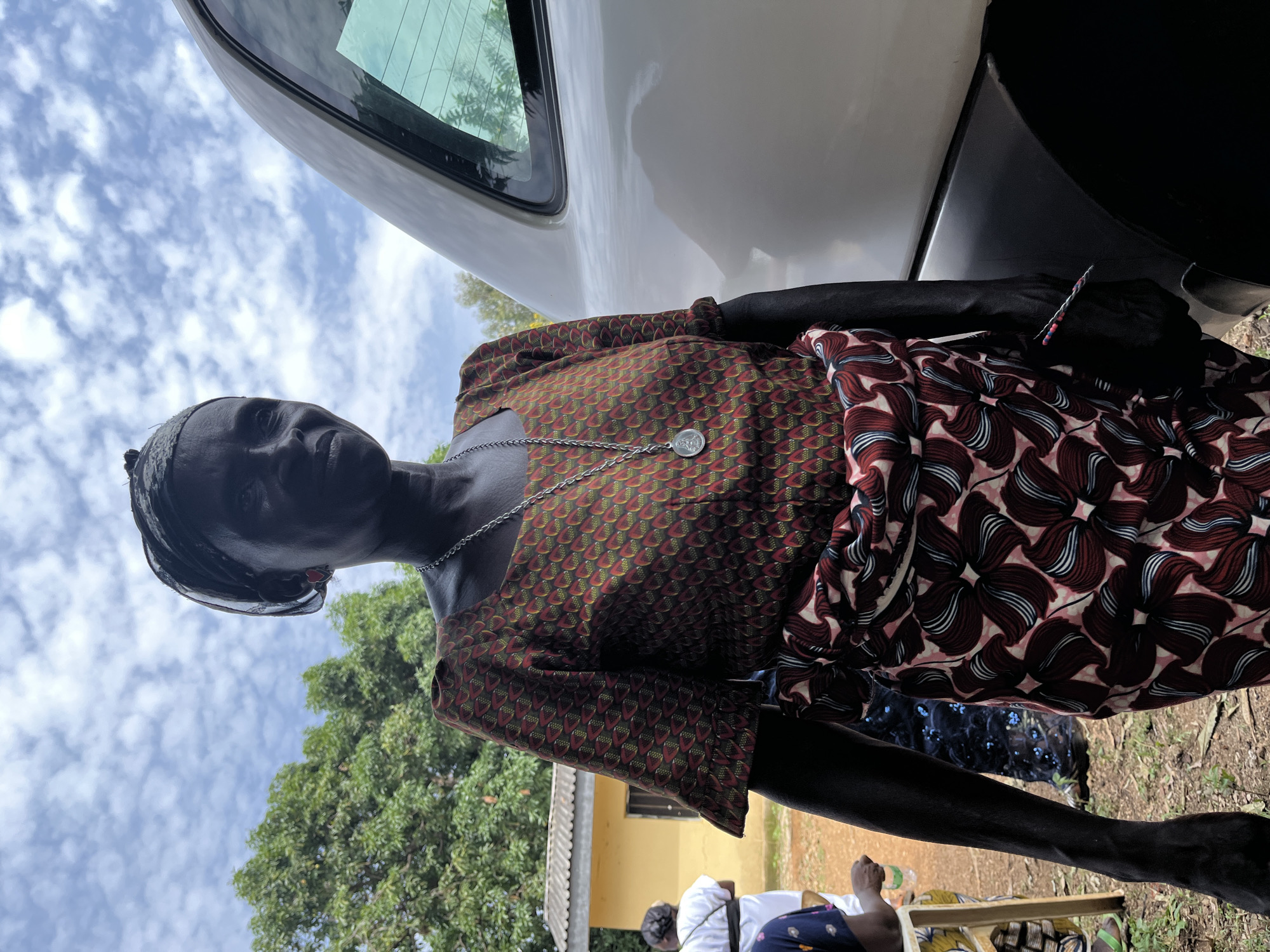 A woman stands next to a white car in Ganawuri, Nigeria. She wears a patterned dress with red and brown geometric designs and a silver necklace. The sky is cloudy, and trees are visible in the background. A yellow building is partially visible behind the woman. Other people are seated nearby, with a glimpse of clothing suggesting a group setting. The ground is covered with vegetation and dirt.