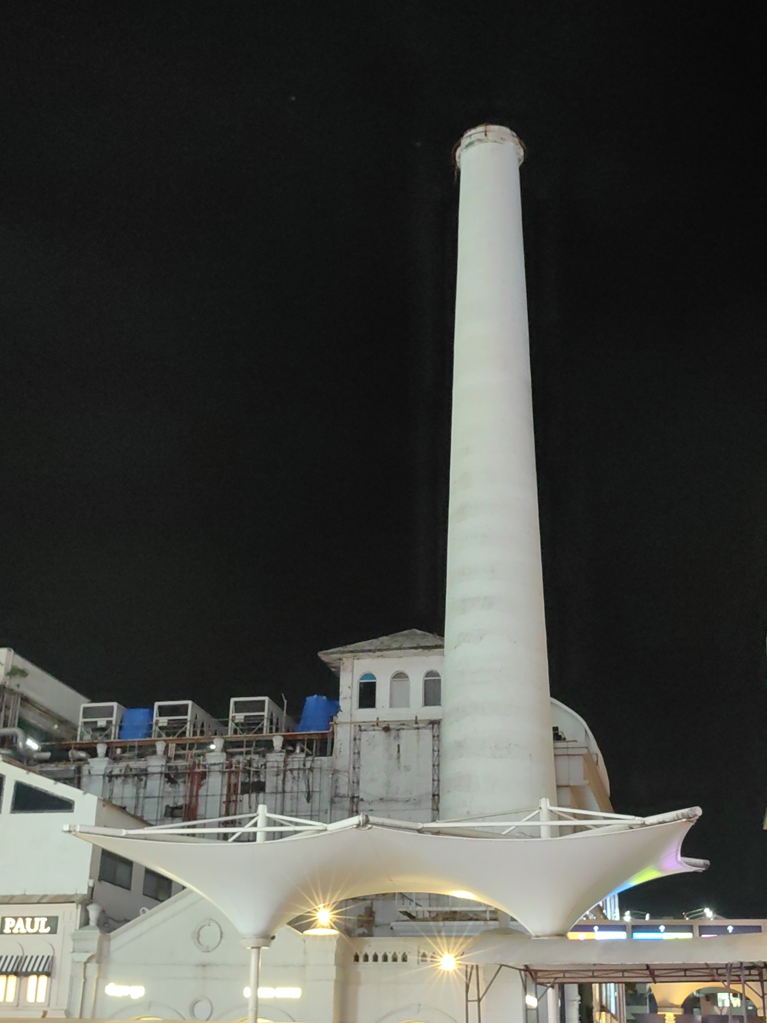 This image captures a striking night view of the iconic Phoenix Marketcity mall in Kurla, Mumbai, India. Dominating the frame is a towering, white cylindrical structure, likely a repurposed industrial chimney, which stands tall against a dark, starless sky. To the left of the chimney, a portion of the mall's facade is visible, featuring a white canopy illuminated from beneath, casting a warm glow. The building exhibits a mix of modern architecture with distinct arched windows and what appear to be industrial air conditioning units on a higher level, some covered with blue tarpaulins. A sign with the word "PAUL" in a clear font is visible on the lower left part of the building. The overall scene suggests a lively urban environment, with the mall's architectural features and lighting indicating it is a popular destination.