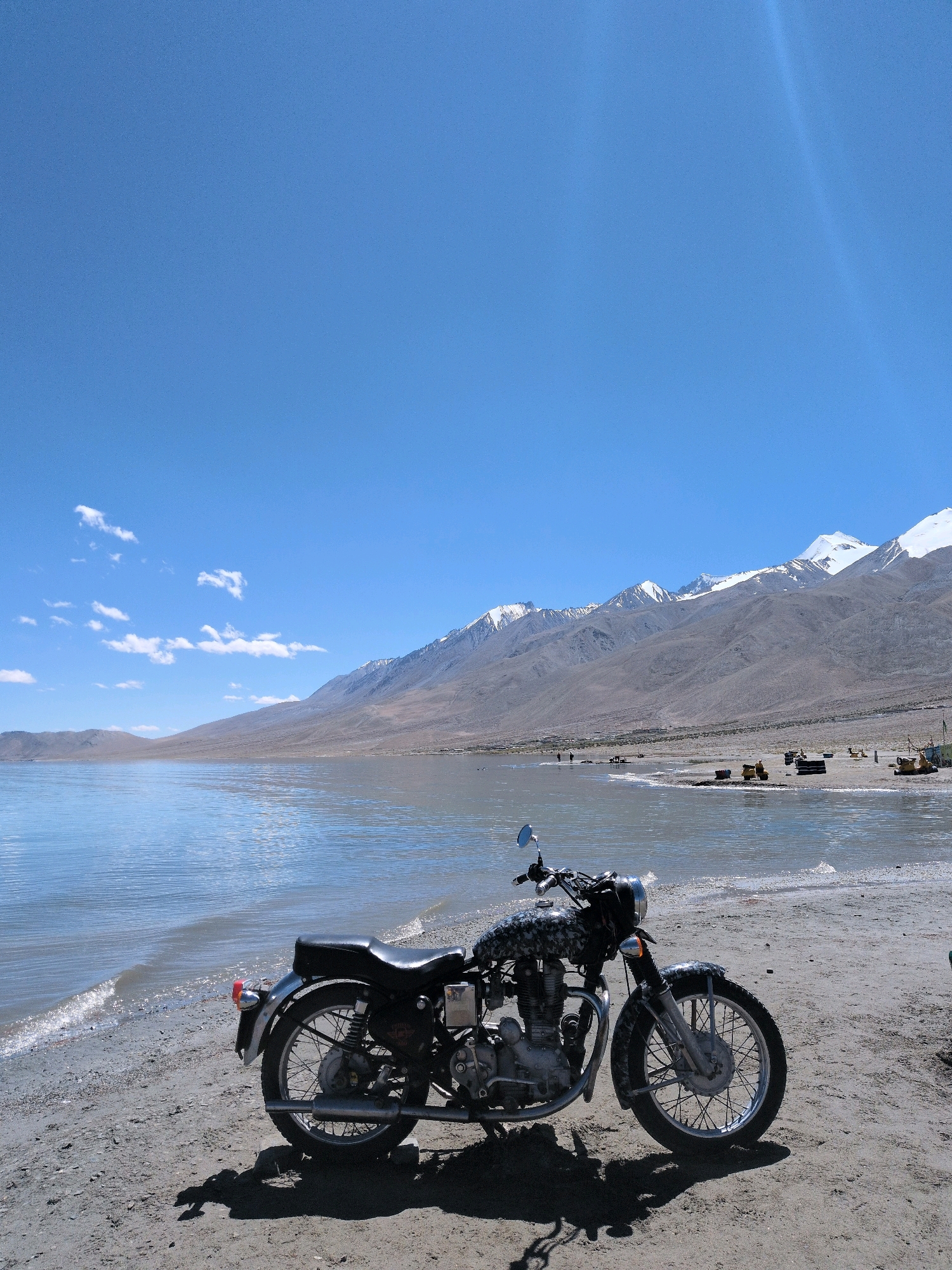 A motorcycle is parked on a sandy shore next to a lake. The motorcycle is black and has a camouflage pattern on the fuel tank. The lake is calm and reflects the blue sky. In the background, there are mountains with snow-capped peaks. There are a few people and some construction equipment visible on the shore in the distance. The sky is clear and blue with a few scattered clouds. The scene appears to be in a natural, remote location.