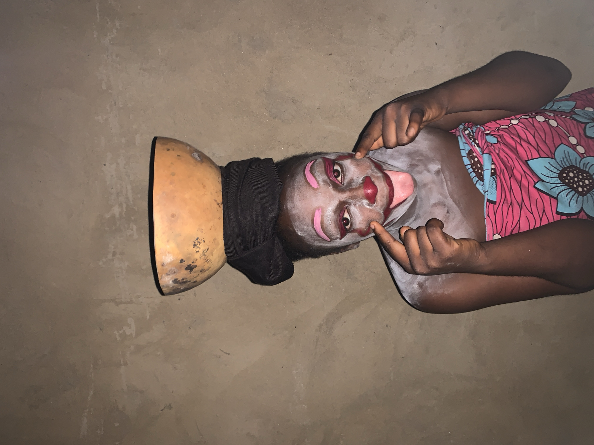 A person with clown-like makeup, including white powder on their face and chest, pink eyebrows, a red nose, and red lipstick, is sticking out their tongue. They are wearing a brightly colored pink and blue floral fabric around their lower body and a black cloth around their head, on top of which rests a large gourd. The background is a textured, light brown wall, consistent with outdoor structures in Agban, Nigeria. The person's fingers are touching their cheeks, emphasizing the drawn-on smile.