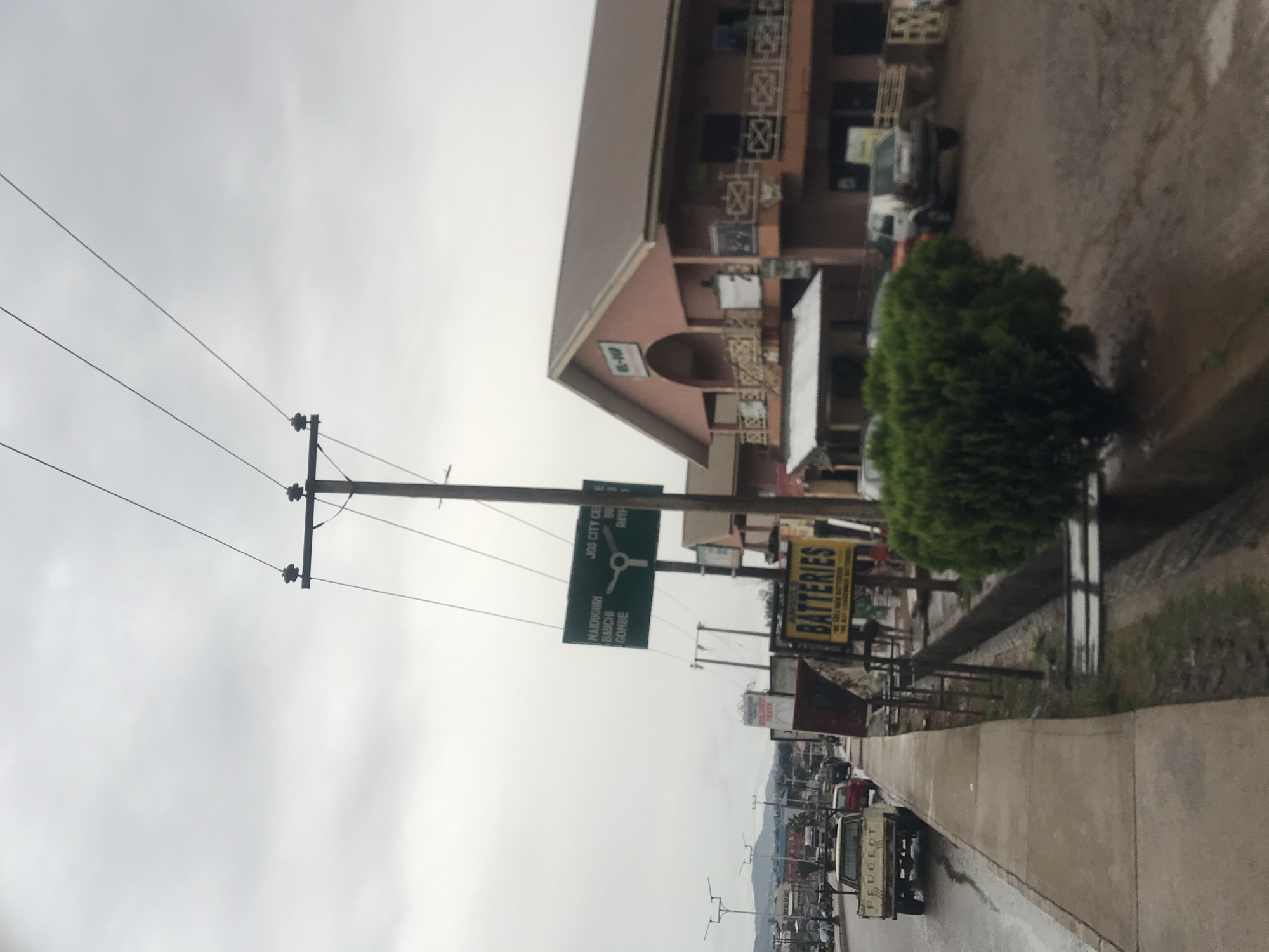 The original image is rotated 90 degrees counter-clockwise, depicting a street scene in Jos, Nigeria, under an overcast sky. The road and sidewalk appear damp, suggesting recent rain.
In the foreground, a prominent utility pole with power lines extends across the top of the frame. To the right of the pole, a large green directional sign is visible. The sign indicates routes to "JOS CITY CENTRE," "MAIDUGURI," "BAUCHI," and "GOMBE," confirming the location context.
Along the right side of the street, there's a sidewalk bordering a dirt verge with a neatly trimmed green shrub. Further down the road, various parked vehicles are visible, including a white Peugeot pickup truck with "PEUGEOT" written on its tailgate.
On the left side of the street, several buildings line the thoroughfare. One multi-story building, painted in a light brown or peach color with intricate white balcony railings, stands out. A sign on this building partially reads "EL-DOP". Adjacent to the directional sign, a large yellow sign advertising "BATTERIES" is clearly visible, with smaller text reading "WE SELL NEW BATTERIES" and "WE BUY USED BATTERIES."
The sky is uniformly grey and cloudy, contributing to the subdued lighting of the scene. No people are discernible in the image, and the overall impression is that of a quiet, everyday street in an urban or semi-urban area.
