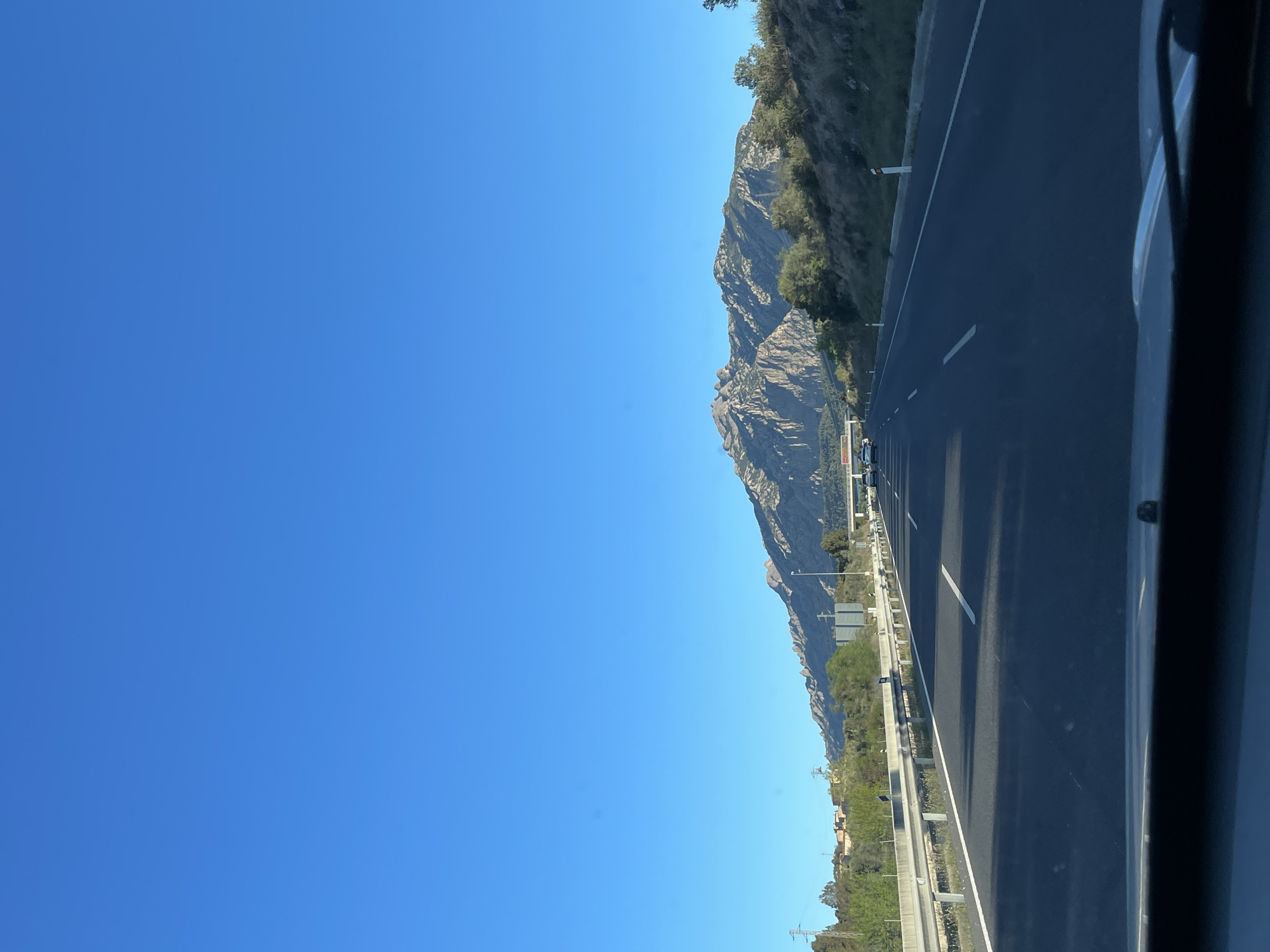 The image captures a view from a vehicle moving on a highway near Esparreguera, Spain. The highway is adjacent to a mountainous region. The sky is clear and blue. The highway is dark asphalt with white lane markings. A metal guardrail separates the road from the mountainside. The mountains are rocky with sparse vegetation.