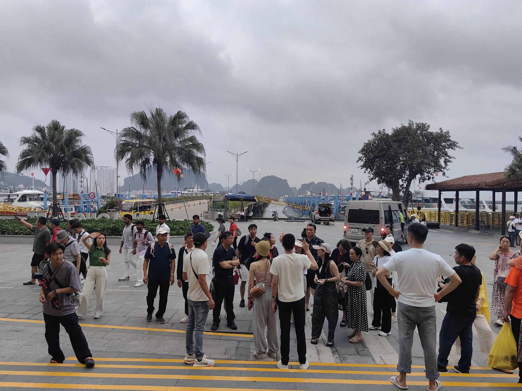 The image shows a group of tourists gathered in Bai Chay Ward, Vietnam. They are standing on a paved area with yellow striped lines, possibly a designated pedestrian zone or a set of steps. Palm trees and other foliage are visible in the background. The sky is overcast, suggesting a cloudy day. Several people are looking around, and some seem to be engaged in conversation or listening to someone. There are vehicles and boats visible in the distance, indicating a waterfront area. The general atmosphere is that of a tourist location, with people observing the surroundings or waiting for transportation.