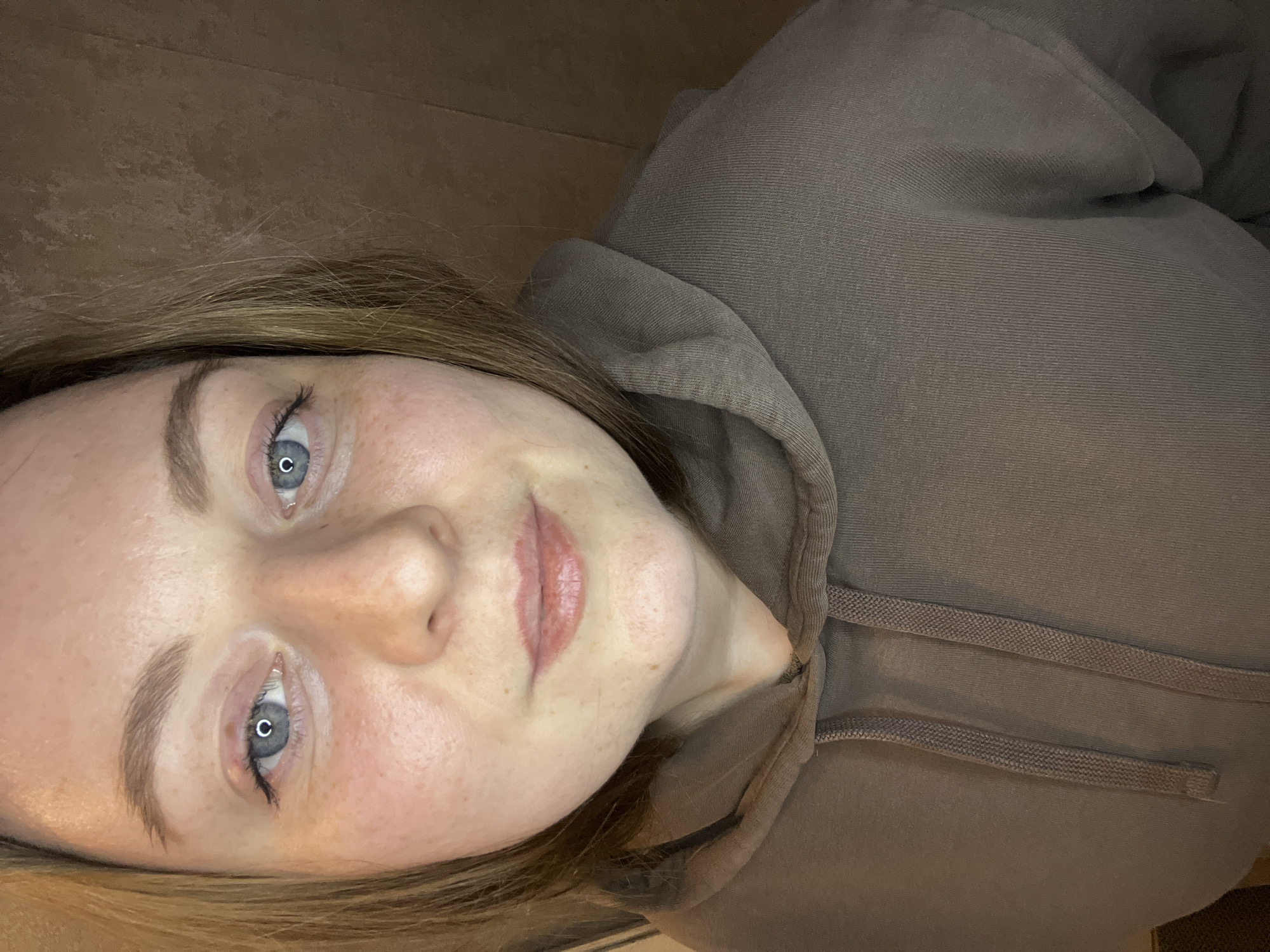 The image shows a woman with fair skin, light brown hair, and blue eyes, wearing a brown hooded sweatshirt. Her face is tilted, and she is looking at the camera. The background is a light brown surface. The location in the city of Раздолье, Russia, cannot be determined from the image.