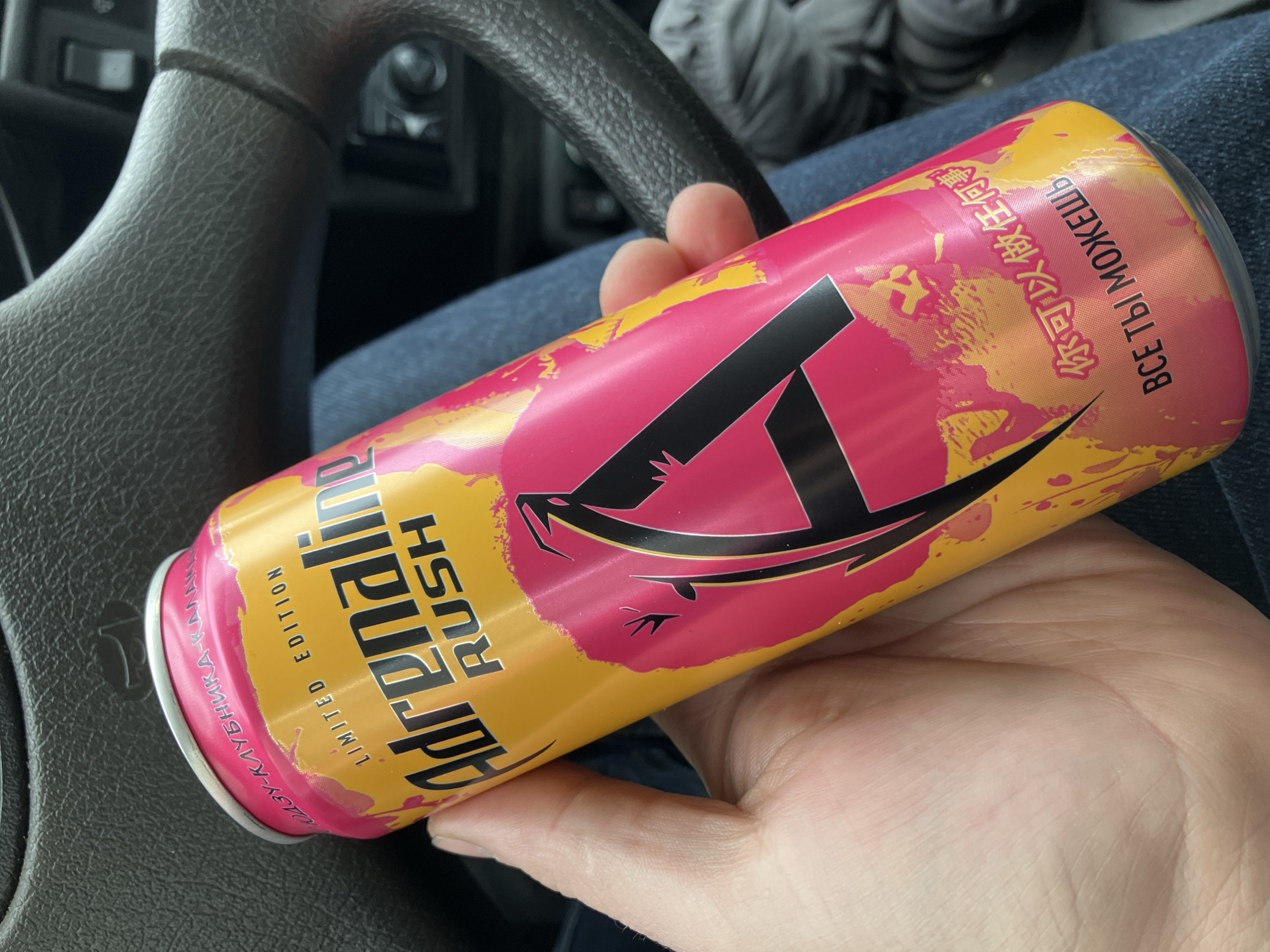 The image shows a person holding a can of Adrenaline Rush energy drink in their hand, likely inside a car given the presence of a steering wheel in the background. The can is a limited edition flavor featuring a pink and yellow design with a black "A" logo. Some text on the can includes "Adrenaline Rush," "Limited Edition," and the flavor name which looks to be related to strawberry. The can also features text in Russian and what appears to be Chinese. The person's hand and part of their blue jeans are also visible. The scene is matter-of-fact, focusing on the presentation of the energy drink. There is no reference to the city of Chaikovsky, Russia, in the image or the visible text.