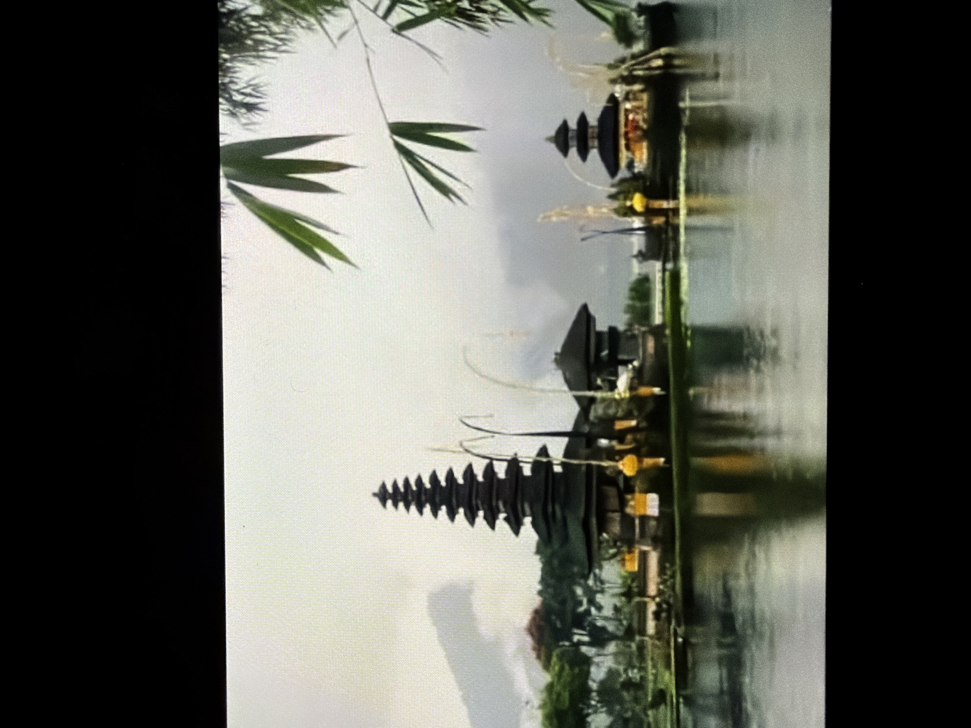 The image shows a landscape in Ungasan, Indonesia. It features two multi-tiered Balinese temples reflected in a body of water. Green foliage and bamboo are visible in the foreground and middle ground. The sky is overcast, suggesting a cloudy day. There are no visible people or explicit actions. The focal point is the architectural structure of the temples and their reflection.