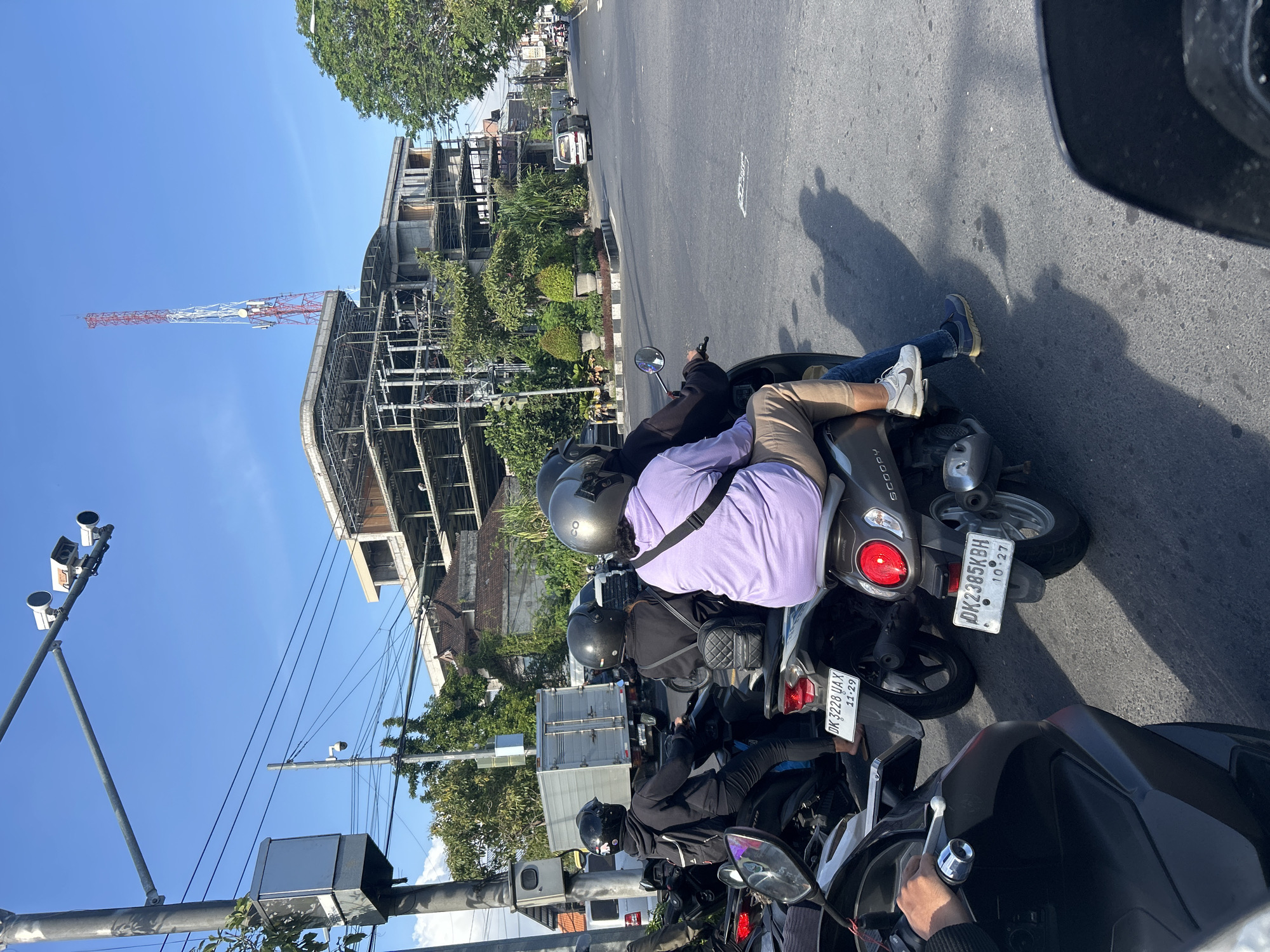 This is an eye-level, medium shot taken on a sunny day in Denpasar, Indonesia, likely on a street with moderate traffic. In the foreground, the rear of a black scooter is visible, with its rider's hand resting on the handlebars. Partially obscured by this scooter is another scooter carrying two passengers wearing helmets. The passenger in the rear wears a light purple hoodie and beige pants, while the driver wears a black hoodie and helmet. Their scooter displays a license plate that reads "DK2385KBH 10 27". Another scooter with two passengers is ahead and to the left. The driver, wearing a black hoodie and helmet, has a backpack. The passenger in the rear is wearing a black hoodie and helmet. This scooter has a license plate reading "DK 3228 UAX 11-29".
Further down the road, traffic is visible, including a white truck with a box trailer, a white van, and other vehicles. A large, unfinished building with exposed concrete and steel beams dominates the background, with a tall red and white telecommunications tower visible behind it. A traffic light with multiple cameras is mounted on a pole on the left side of the road. The sky is clear and blue. The environment appears to be an urban street with trees and some greenery.