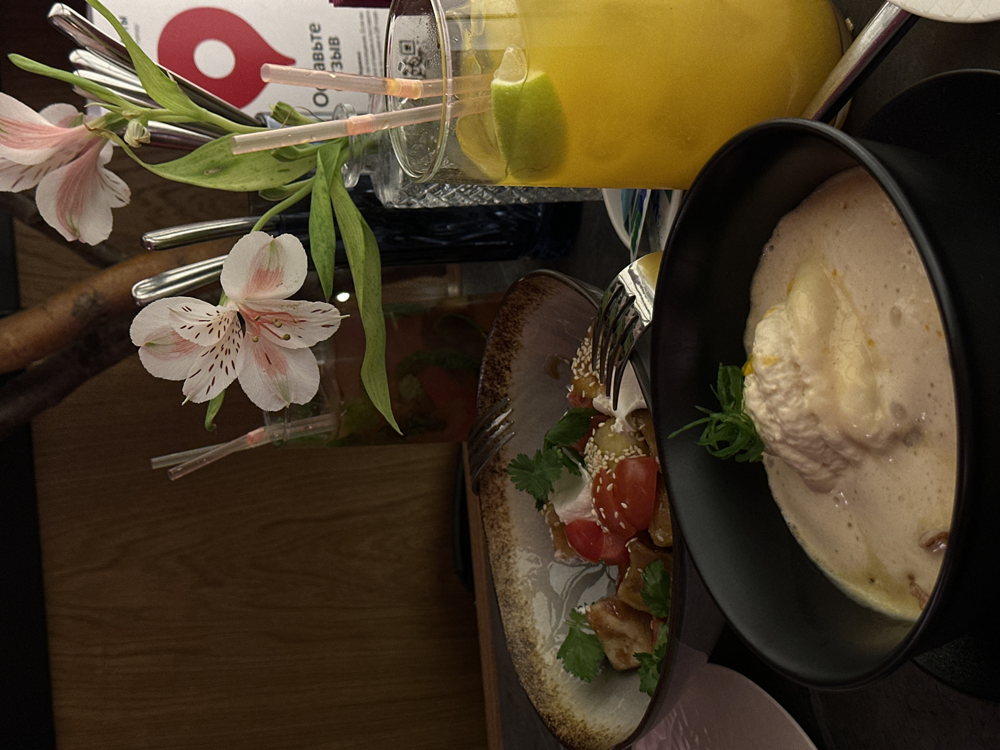 The image shows a dining table with several items of food and drink. A tall glass of yellow liquid with a lime wedge sits at the top of the frame. Below it is a plate with a salad of tomatoes, sesame seeds, and herbs, with a fork partially piercing the salad. Next to this plate is a black bowl containing a creamy soup with a dollop of some kind of topping, garnished with green herbs. A flower arrangement with pink and white alstroemeria flowers and green foliage is also visible on the table. The environment appears to be a restaurant setting, with a wooden table surface. The location, as mentioned, is Ovoshchnoy, Russia, though this detail cannot be definitively confirmed from the image content alone.