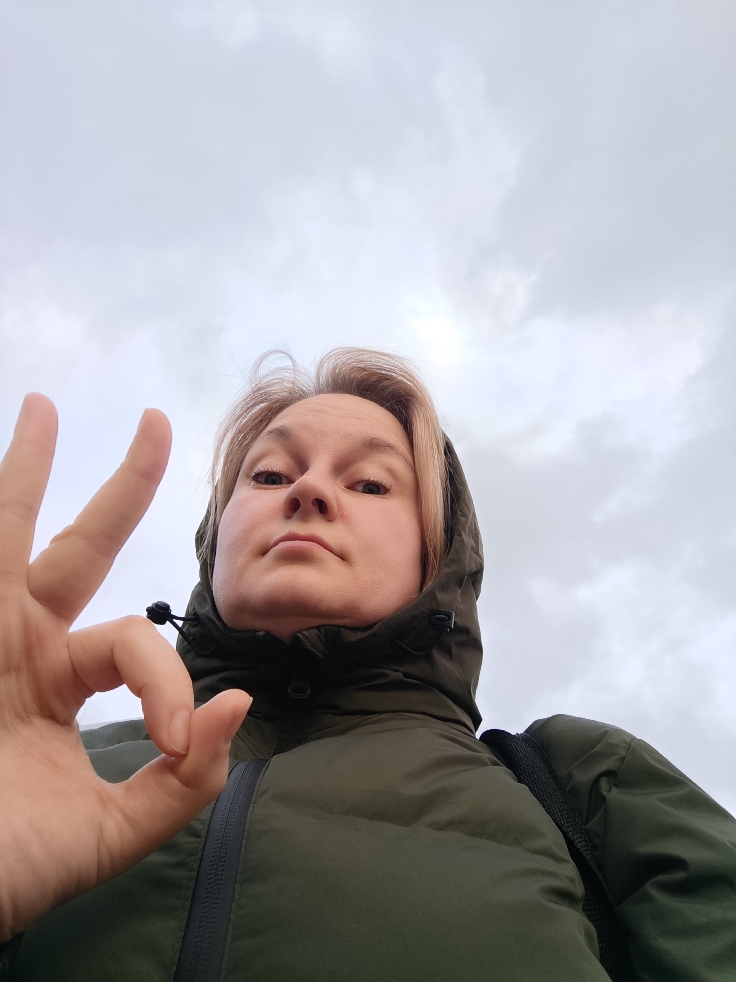 A close-up, low-angle shot features a person making an "OK" hand gesture against an overcast sky. The subject, likely female, has light blonde hair and is wearing a dark green hooded winter jacket or coat, with the hood up around her head. Her left hand is prominent in the foreground, forming the "OK" sign with the thumb and index finger touching, while the other fingers are extended upwards. Her face is directed towards the camera, displaying a neutral to slightly serious expression. A black strap, possibly from a backpack, is visible over her right shoulder. The background is a pale, cloudy sky, indicating daytime and overcast weather. No text is visible in the image. The location context for this image is Markovskii, Russia.