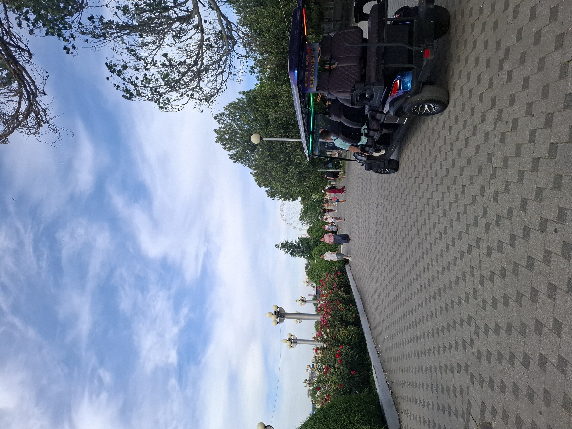 A golf cart is parked on a paved walkway in a park-like setting under a blue sky with white clouds. The golf cart is dark-colored with multiple rows of seats and has neon lights along its roof. Several people are walking on the walkway in the background. To the left of the walkway, there is a flower bed with red and white roses, followed by manicured bushes and lampposts. In the distance, a Ferris wheel is visible. Trees line the right side of the walkway, with branches extending over the top of the image. The walkway is paved with gray interlocking bricks.