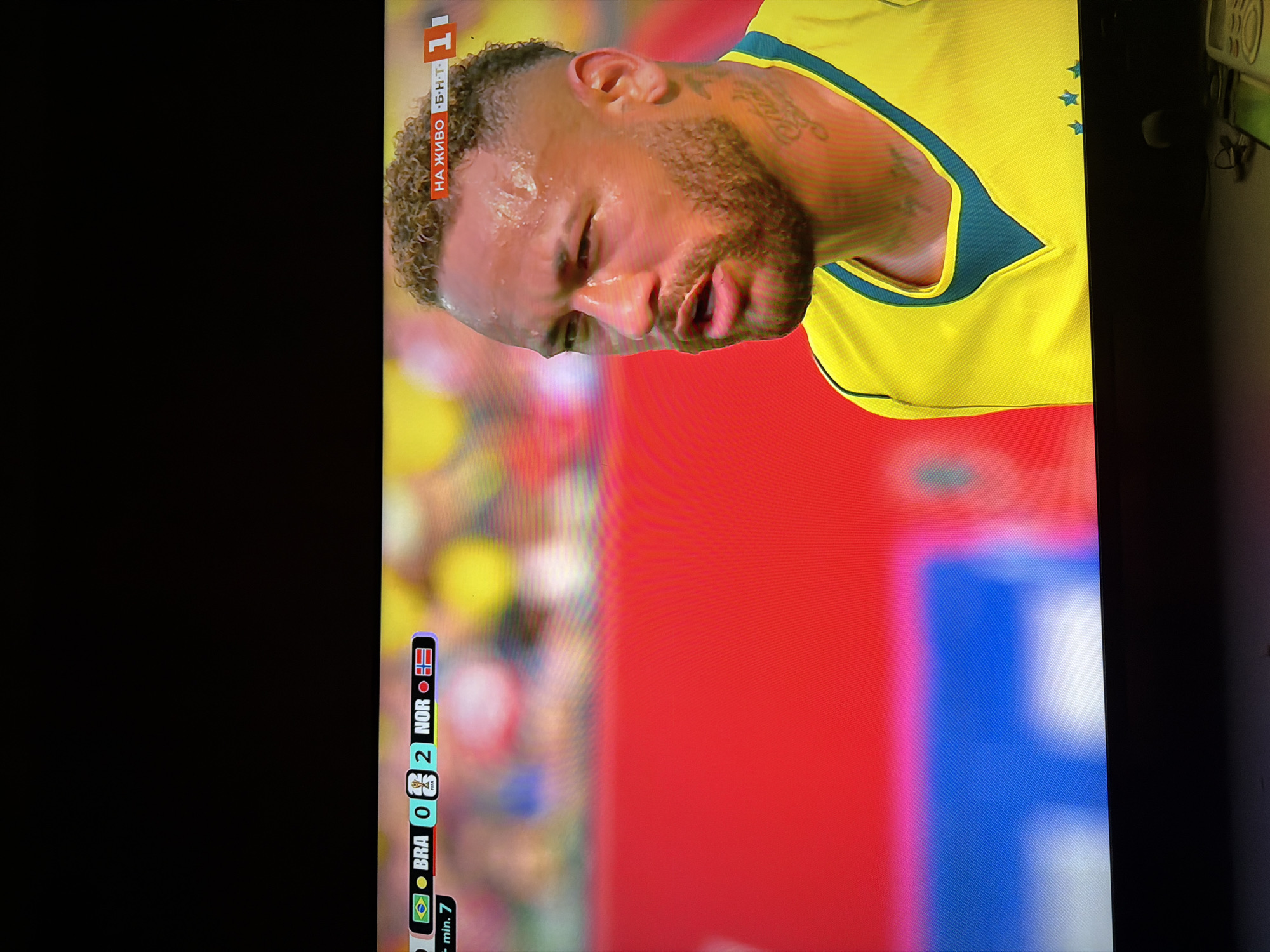 The image is a close-up of a television screen displaying a soccer match. In the foreground, on the right, is a close-up of the face and upper torso of a soccer player, identified by his yellow jersey with blue trim and three stars. The player has short, curly dark hair, a beard, and is sweating. He is looking off to his right. In the lower left corner of the screen, a score graphic shows "BRA 0 2 NOR," indicating Brazil is playing Norway, and Brazil has 0 goals while Norway has 2. A timer shows "- min. 7". To the right of the score, there is a graphic that says "НА ЖИВО Б•Н•Т 1". The background of the soccer match is blurred, showing spectators and a field. The television screen is positioned in front of a dark surface, likely furniture, and a remote control is visible on this surface in the lower right corner of the image. The image quality suggests it was taken with a handheld device, possibly a phone, and there is a slight distortion or reflection on the screen. There is no discernible connection to the city of Ognianovo, Bulgaria, within the image itself.