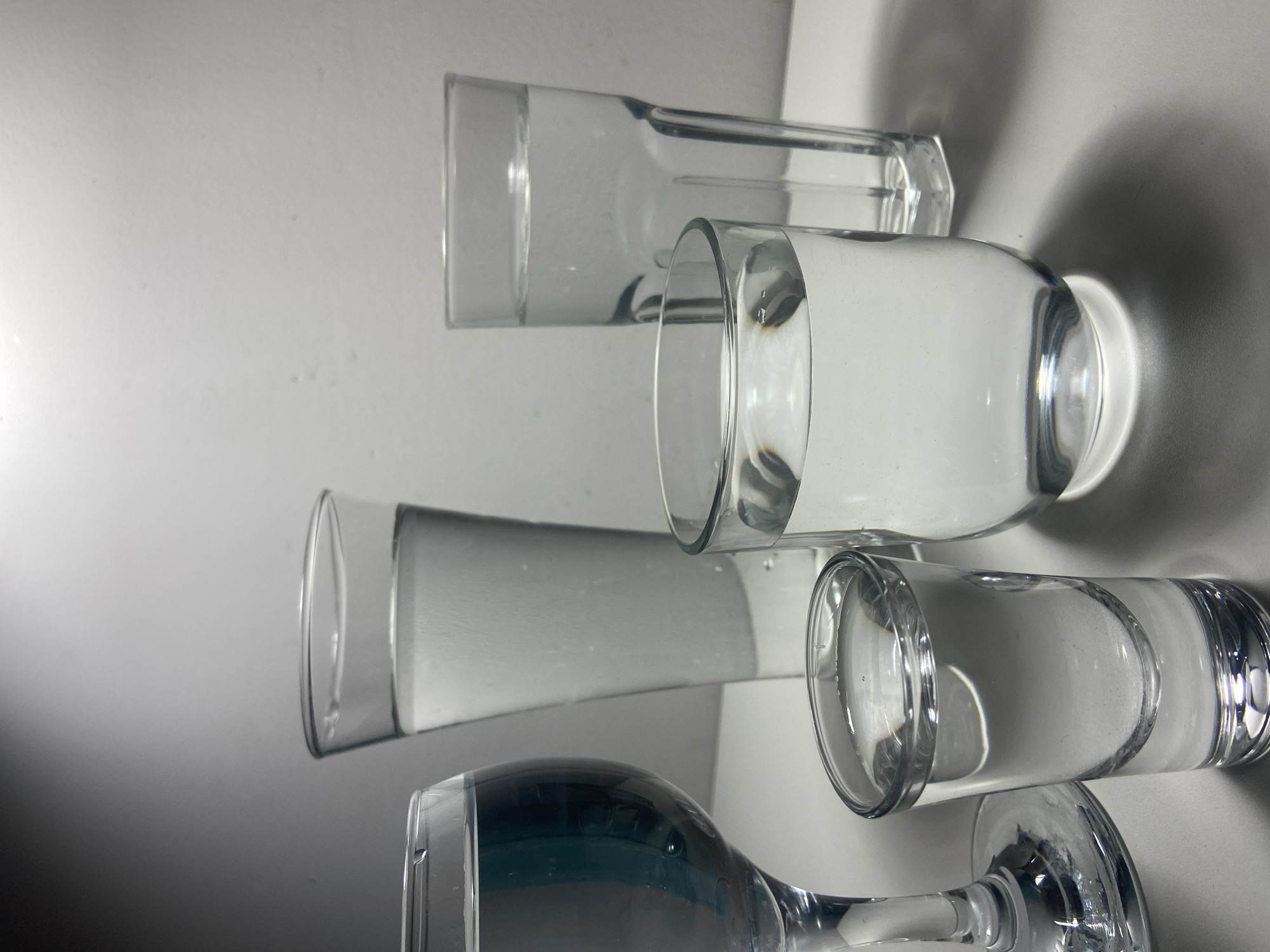 The scene, presented as being located in the city of Budva, Montenegro, depicts a still life arrangement of five transparent glass vessels containing clear liquid, consistent with water, on a plain, light-colored surface. A uniform, light-colored wall serves as the background.
From left to right:
1. A stemmed wine glass, partially visible on the left, is filled with clear liquid to approximately two-thirds of its capacity. A dark vertical reflection is visible within its bowl.
2. In the immediate foreground, a small, cylindrical shot glass is filled with clear liquid, nearly to the brim.
3. Behind the shot glass, a tall, slender, slightly conical glass is filled with clear liquid, exhibiting small air bubbles clinging to its interior surface, reaching approximately three-quarters full.
4. To the right and slightly in front of the tall conical glass, a shorter, wider, rounded tumbler-style glass holds clear liquid, filled to about two-thirds of its volume.
5. Furthest to the right, a tall, rectangular-shaped glass with subtly paneled sides contains clear liquid, also displaying small air bubbles on its interior, filled to approximately three-quarters.
The surface beneath the glasses is smooth and reflective, casting distinct shadows from each vessel towards the lower right, consistent with a light source positioned above and to the upper left. No individuals, actions, or interactions are present in the image.
