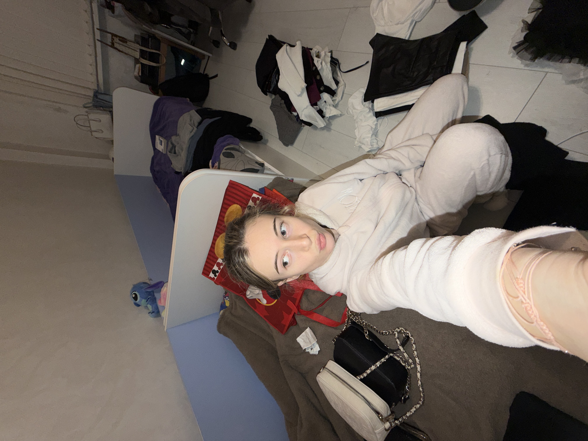 A young female, appearing to be in her late teens or early twenties, is depicted taking a selfie in what appears to be a bedroom located in Plovdiv, Bulgaria. She has light-colored hair and fair skin, and is wearing a light-toned, possibly off-white or light pink, fleece two-piece loungewear set consisting of a hooded top and pants. Her right arm is extended towards the camera, revealing a light pink string bracelet on her wrist. Her expression features a slight pout, and her eyes are directed upwards and to the right.
The woman is seated or kneeling on a dark brown, textured blanket which covers a bed on the left side of the frame. Two handbags are positioned on this blanket next to her: a white quilted rectangular bag with a silver chain strap and a black rectangular bag also with a silver chain strap. Crumpled white paper is visible on the blanket near the handbags.
Behind the subject, a bed with a white headboard and light blue side paneling is visible, covered with a purple blanket. A small blue plush toy resembling Disney's Stitch rests on top of the blue paneling. Further back, on what appears to be another shelf or bed frame, a white handbag and a light blue bag are present.
The floor on the right side of the image is light-colored, possibly laminate or tile. Multiple piles of clothes are scattered across the floor, including a substantial heap of assorted garments (whites, grays, darks) to the subject's immediate right, and a separate pile of dark clothing featuring a black leather-like top or corset and white garments further to the right. A sheer black garment, possibly tulle, is visible near the bottom right corner. In the background, a dark office chair with five caster wheels, a wooden easel or stand, and shelves with various items contribute to the cluttered environment. The room has light-colored walls, with a section of the ceiling appearing sloped.