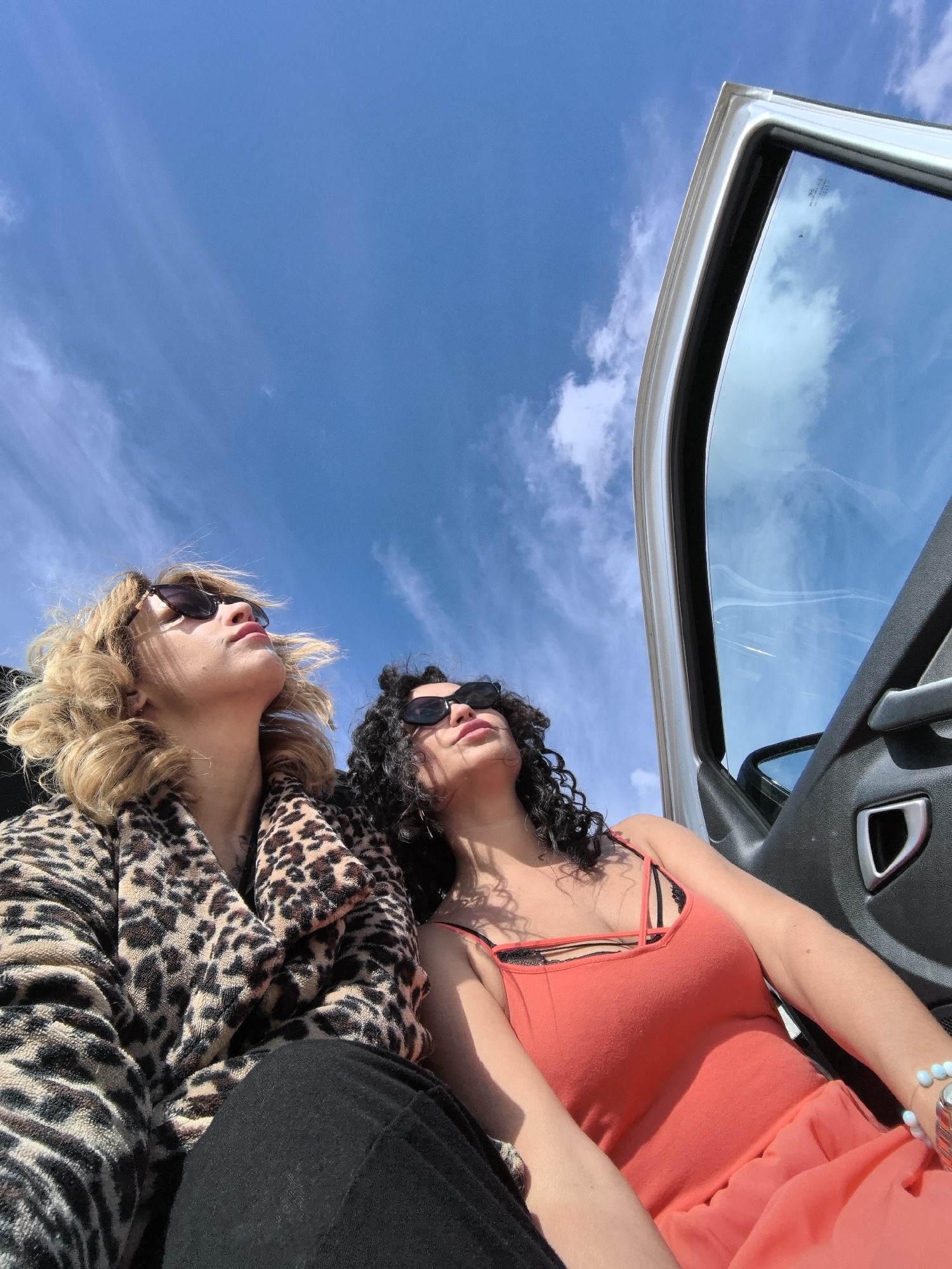 Two women are captured from a low-angle perspective, seated within a car with its passenger door open, looking upwards towards a bright blue sky with sparse, wispy white clouds. The setting appears to be outdoors and sunny, indicative of daytime.
The woman on the left has blonde, curly hair and wears dark sunglasses, a brown and black leopard print coat or robe, and black trousers. Her head is tilted back, with her chin slightly raised.
The woman on the right has dark, curly hair and also wears dark sunglasses. She is dressed in an orange or coral-colored sleeveless top, revealing a black lacy bralette underneath. A light blue beaded bracelet is visible on her left wrist. Her head is similarly tilted back, gazing upward.
The car's white door frame and dark interior panel with a silver handle are visible on the right side of the image, reflecting the blue sky and clouds in the window glass. There is some unreadable text or etching on the car window. The overall mood is relaxed, suggesting the women are enjoying the weather or view. The location context provided is Chaikovsky, Russia.