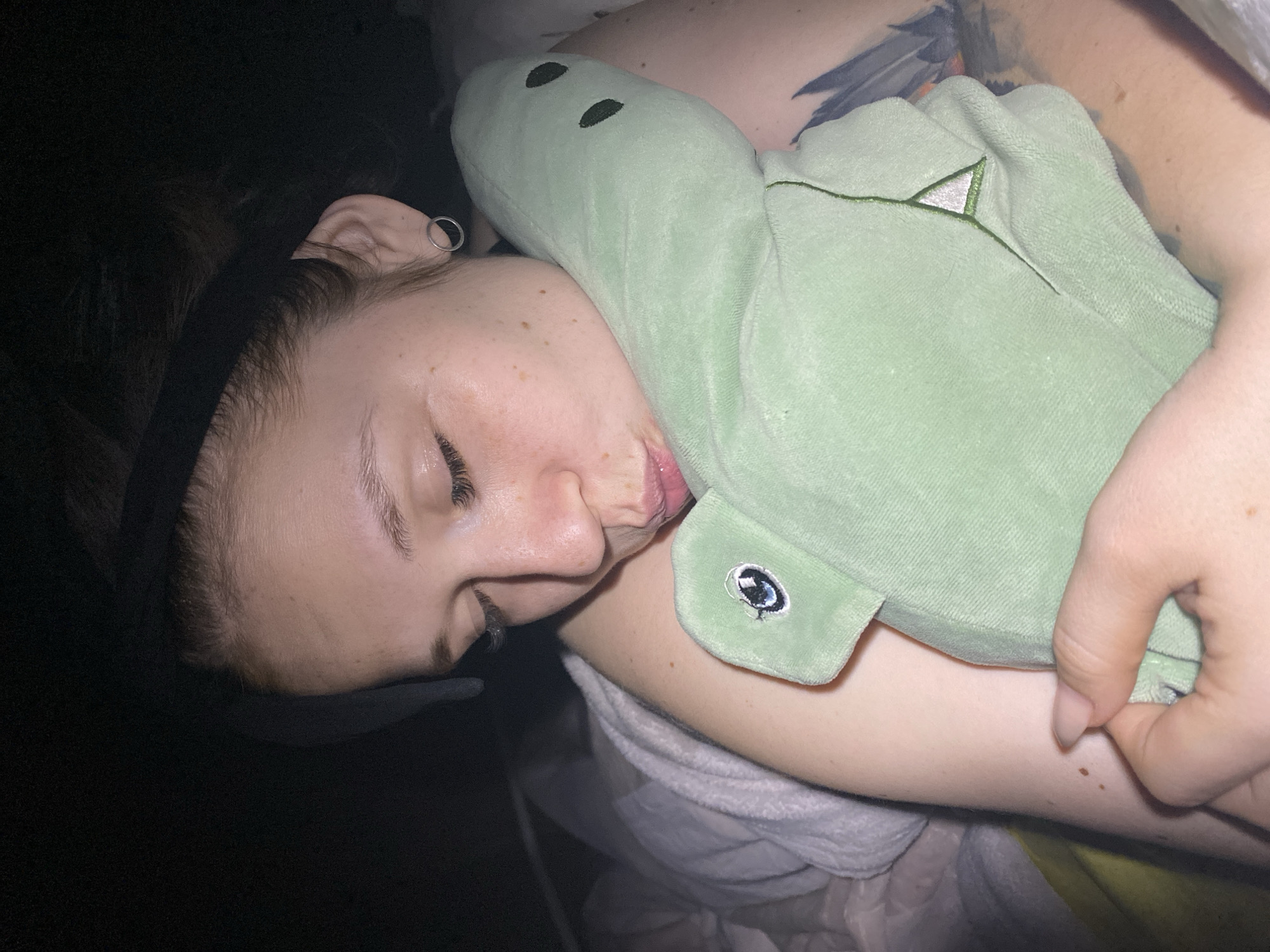 A woman is sleeping on her left side, hugging a light green plush dinosaur. She is wearing a black headband. Her left ear is visible, with a silver hoop earring. The woman has fair skin with freckles. A tattoo is visible on her upper chest. The background is dark and out of focus. There is no evidence to indicate the location in Chaikovsky, Russia.