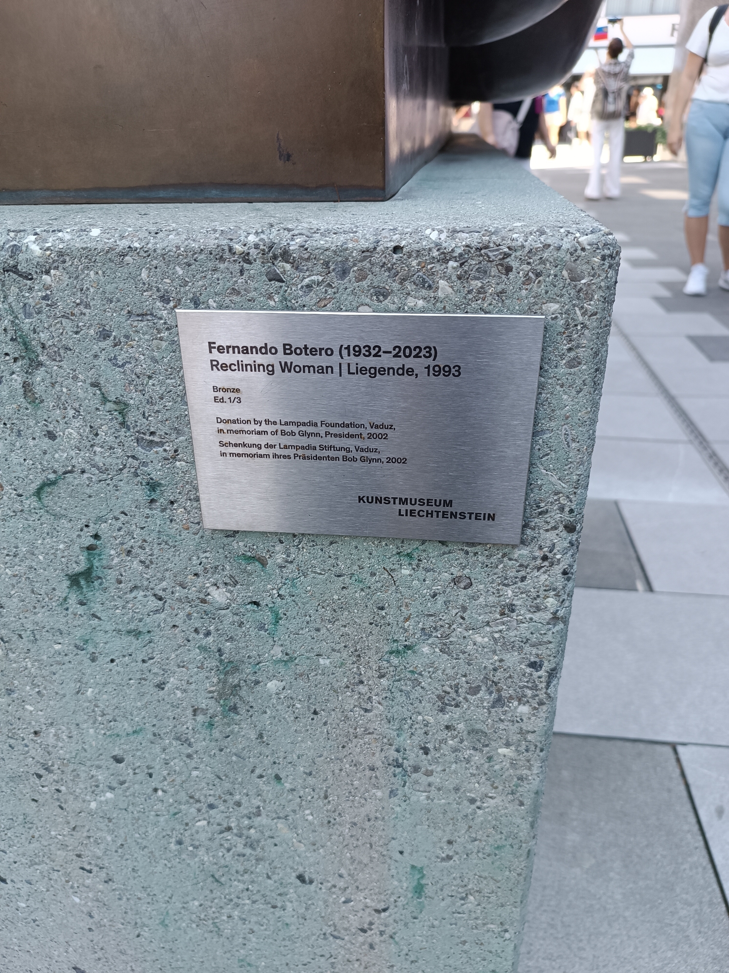 The image shows a metallic plaque attached to a textured concrete pedestal. The plaque reads: "Fernando Botero (1932-2023) Reclining Woman | Liegende, 1993. Bronze Ed. 1/3. Donation by the Lampadia Foundation, Vaduz, in memoriam of Bob Glynn, President, 2002. Schenkung der Lampadia Stiftung, Vaduz, in memoriam ihres Präsidenten Bob Glynn, 2002. KUNSTMUSEUM LIECHTENSTEIN." The background is blurred and shows people walking in an outdoor urban setting with paved ground. The plaque indicates the artwork is located in Vaduz, Liechtenstein, and is part of the Kunstmuseum Liechtenstein.