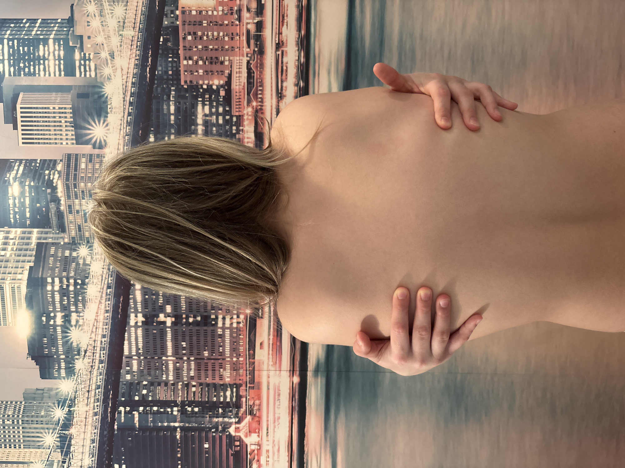 A nude woman is depicted from the back, with her hands resting on her upper back and shoulders. Her head is turned to the left. She has blonde hair with highlights. The background features a blurred, abstract representation of a city skyline, overlaid with an undefined water body. The image appears to be rotated, as the background city scene is vertical while the woman is positioned horizontally.