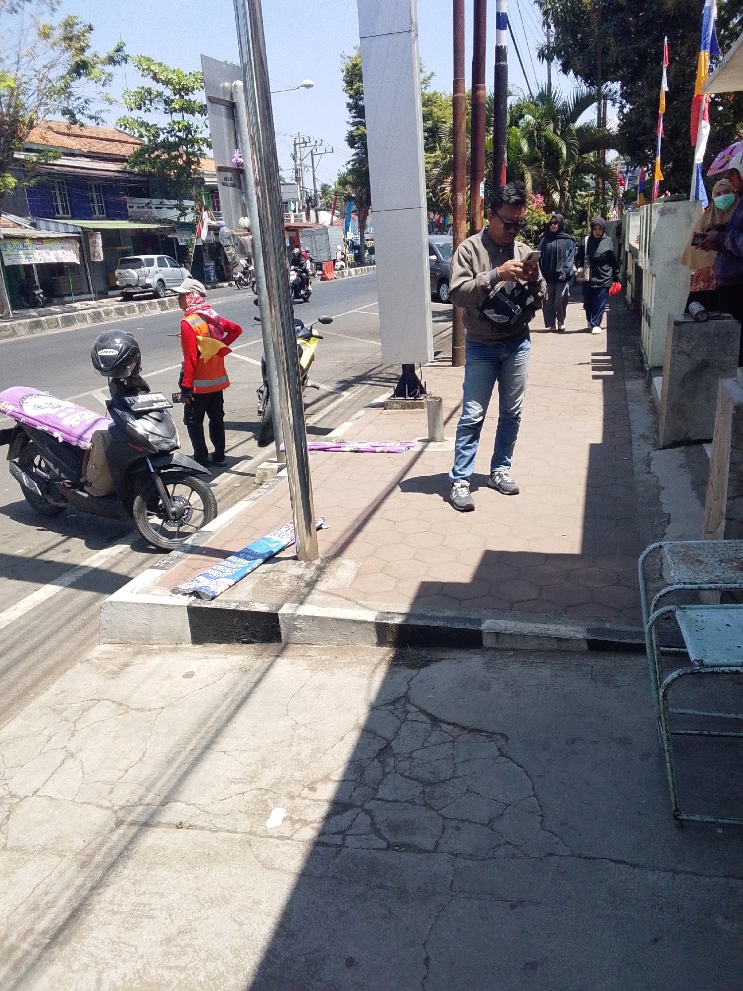 This is a street scene in Purworejo, Indonesia. A person in a red and orange vest stands on the sidewalk next to a parked motorcycle. The motorcycle has a purple cover on its seat. Across the street, a white SUV is parked in front of a building with blue walls. Other pedestrians are visible on the sidewalk in the background. The scene is illuminated by bright sunlight, casting shadows on the sidewalk and street.