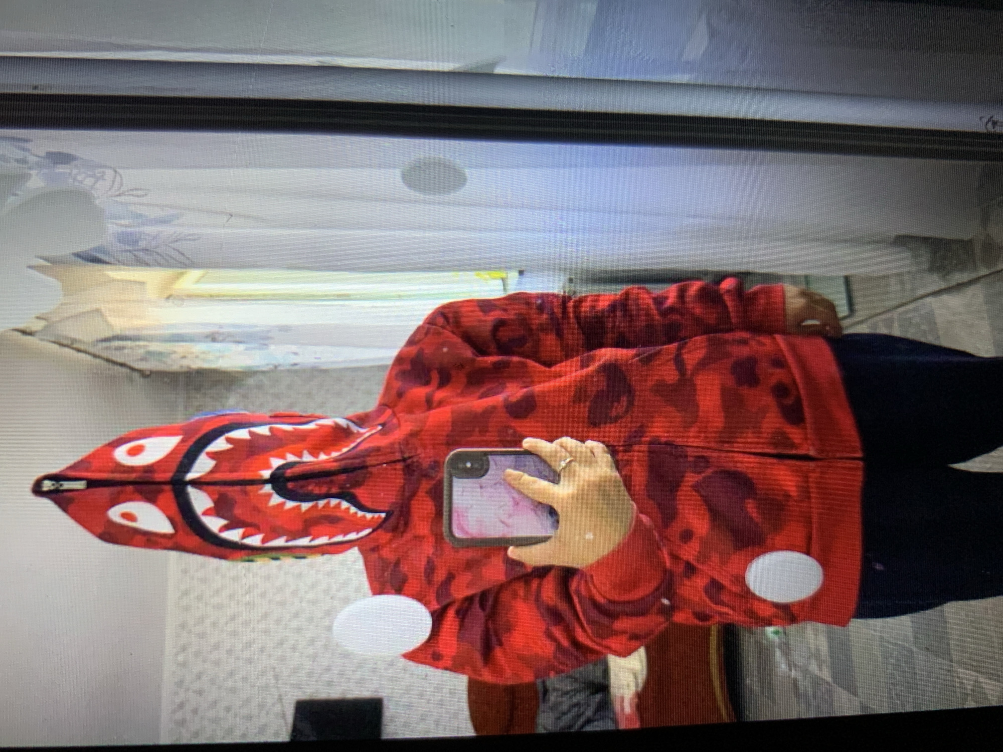 The image shows a person wearing a red Bape hoodie with a shark face design on the hood. The person is holding a phone in their right hand, and there is a ring on their left hand. The background is blurry but appears to be an indoor setting with a mirror and curtains. There are two white circular stickers on the hoodie. The image is a photograph taken in Lymanka, Ukraine.