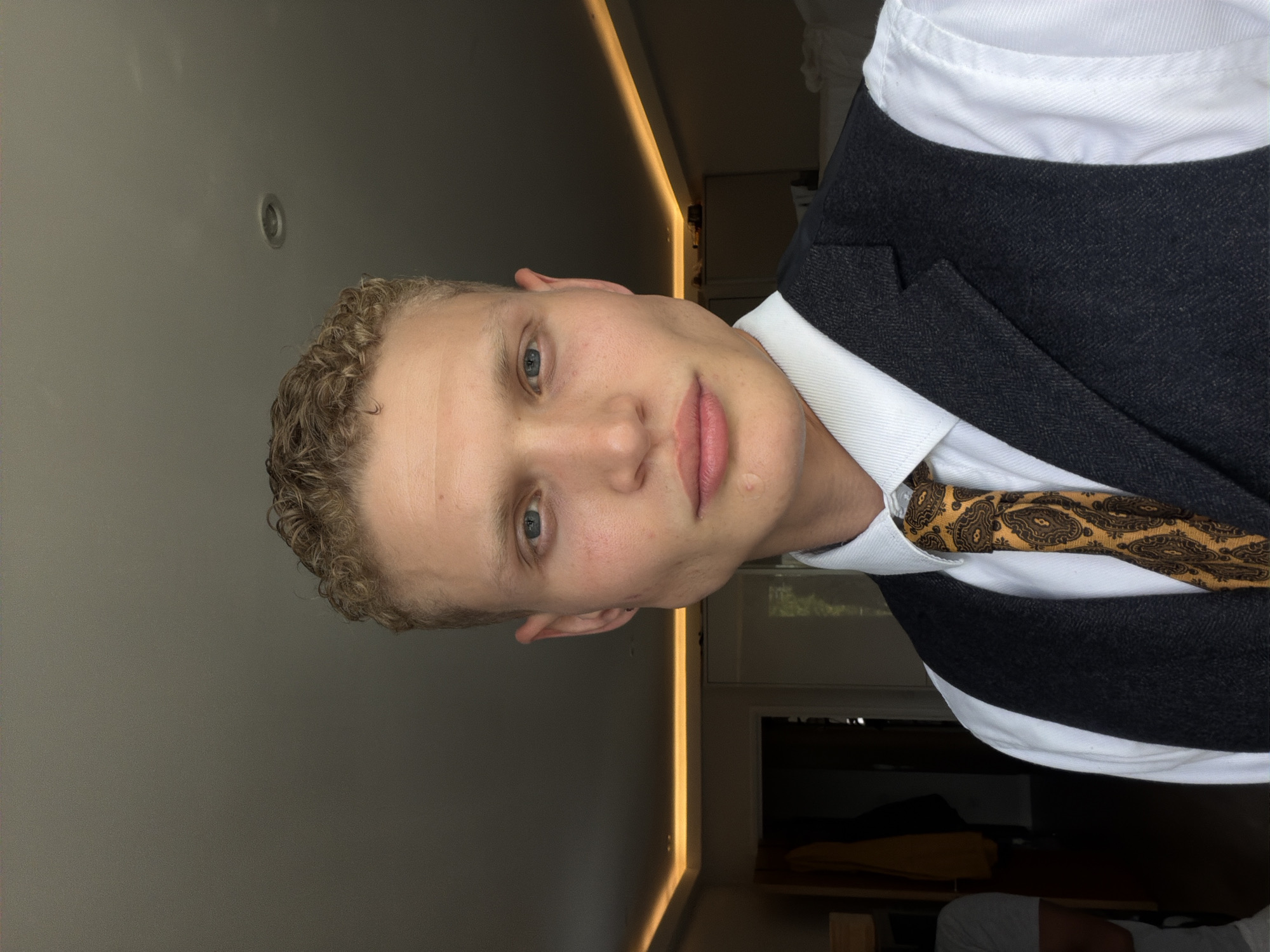 A medium shot captures a fair-skinned, light-haired young man with blue eyes from the chest up, looking directly at the camera. He has a fair complexion, short curly blonde hair, and prominent cheekbones. He is wearing a white collared shirt, a dark grey tweed waistcoat, and a patterned tie with yellow and dark brown paisley designs. The background is dimly lit, suggesting an indoor setting, possibly a bedroom. A strip of warm yellow LED lighting runs along the ceiling edge. The man appears to be standing in a room in London, United Kingdom.