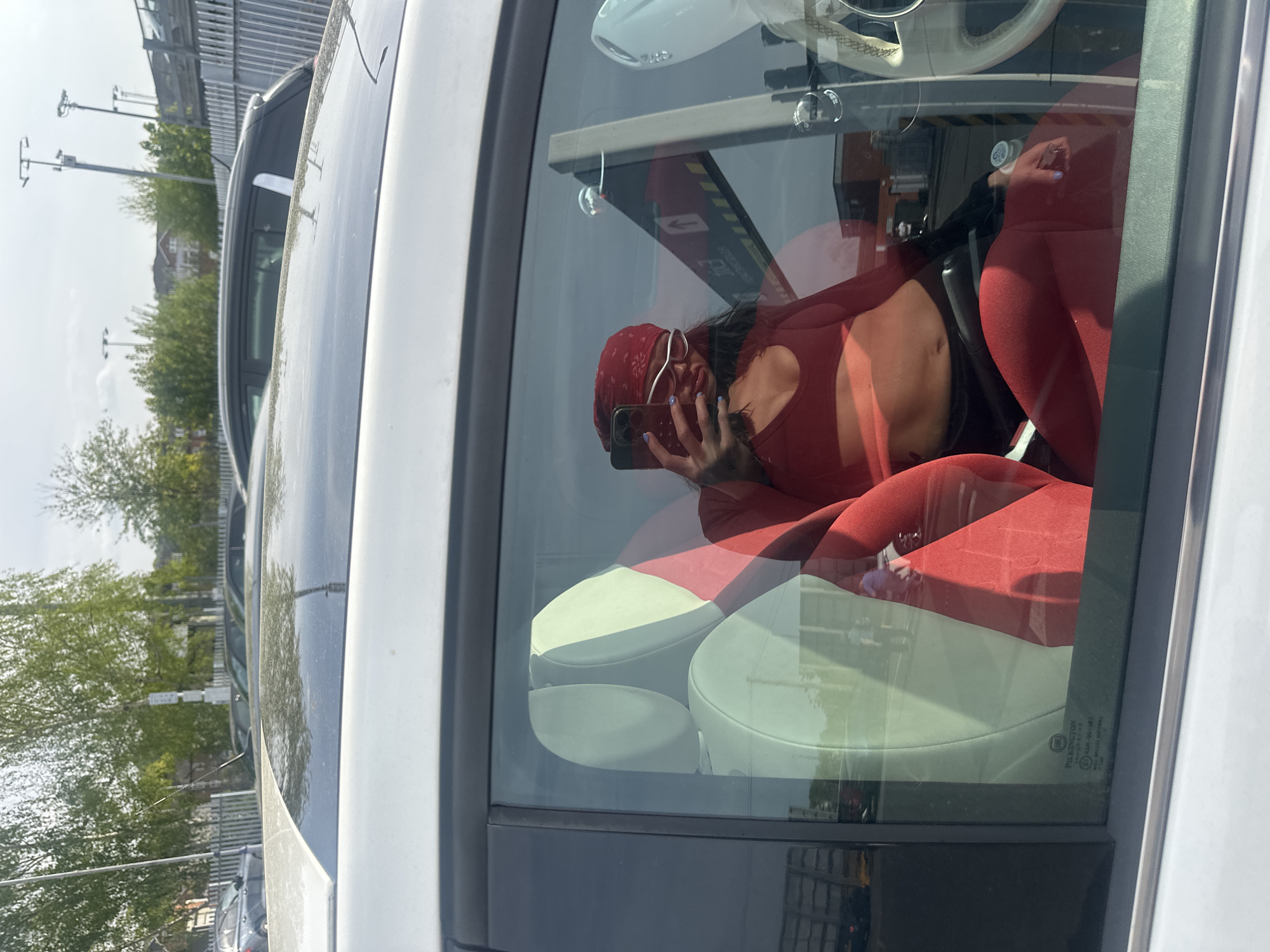 A woman wearing a red bandanna and red top is sitting inside a white car in what appears to be a parking lot. She is holding a phone and taking a picture of herself in the reflection of the car window. The car has red and white seats and a white steering wheel. Outside the car window, there are trees and utility poles in the background, along with buildings that suggest an urban environment in Sevenoaks, United Kingdom.