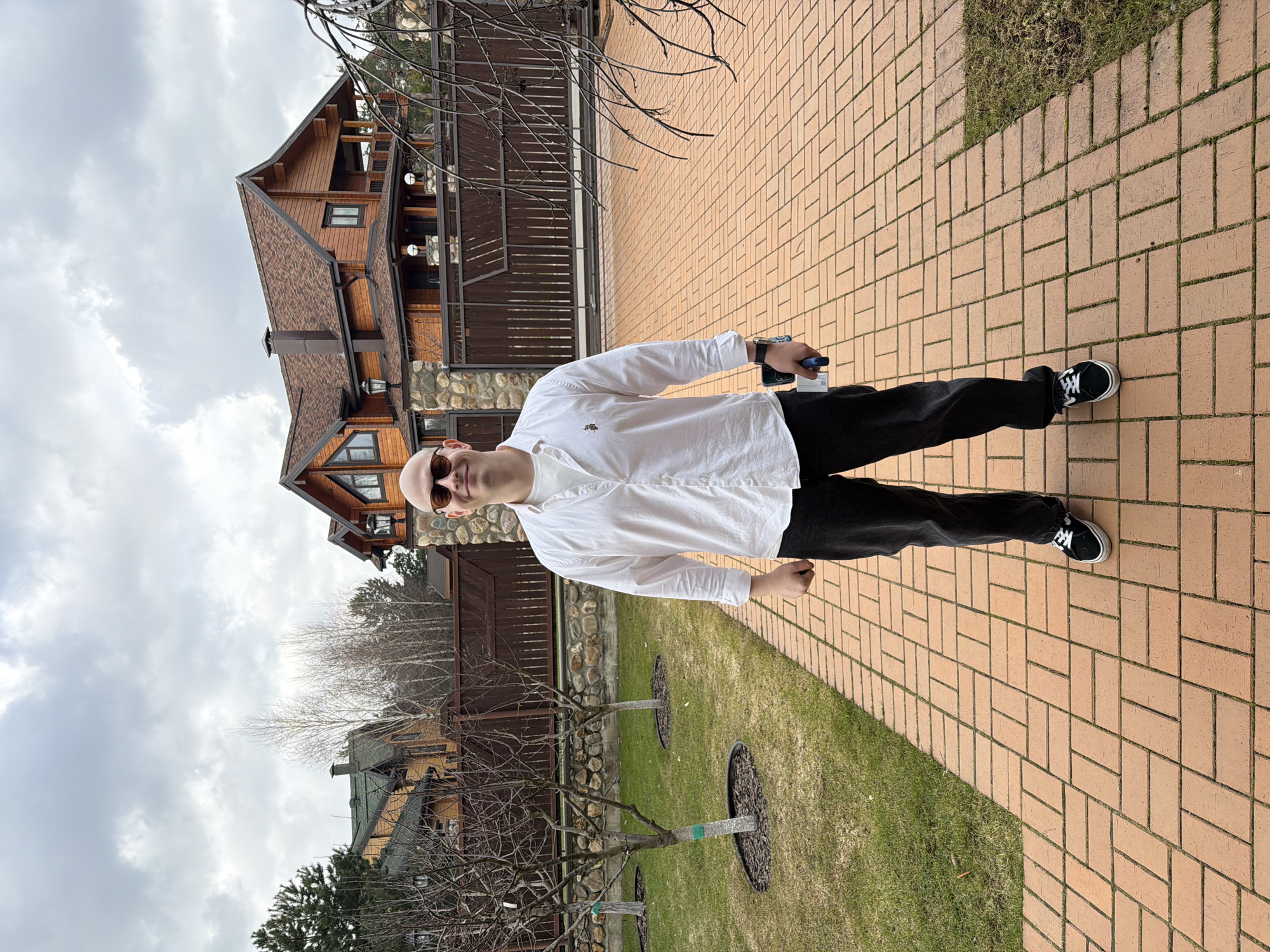 A bald man with sunglasses stands on a brick walkway in front of a house and a green lawn. He wears a white, long-sleeved shirt and dark pants with black and white shoes. He holds a smartphone and a small, blue object in his left hand. The house is a two-story wooden structure with a brown roof and stone accents. A wooden fence runs along the property line. Bare trees are visible in the yard. The sky is cloudy. Mishutkino, Russia cannot be confirmed.