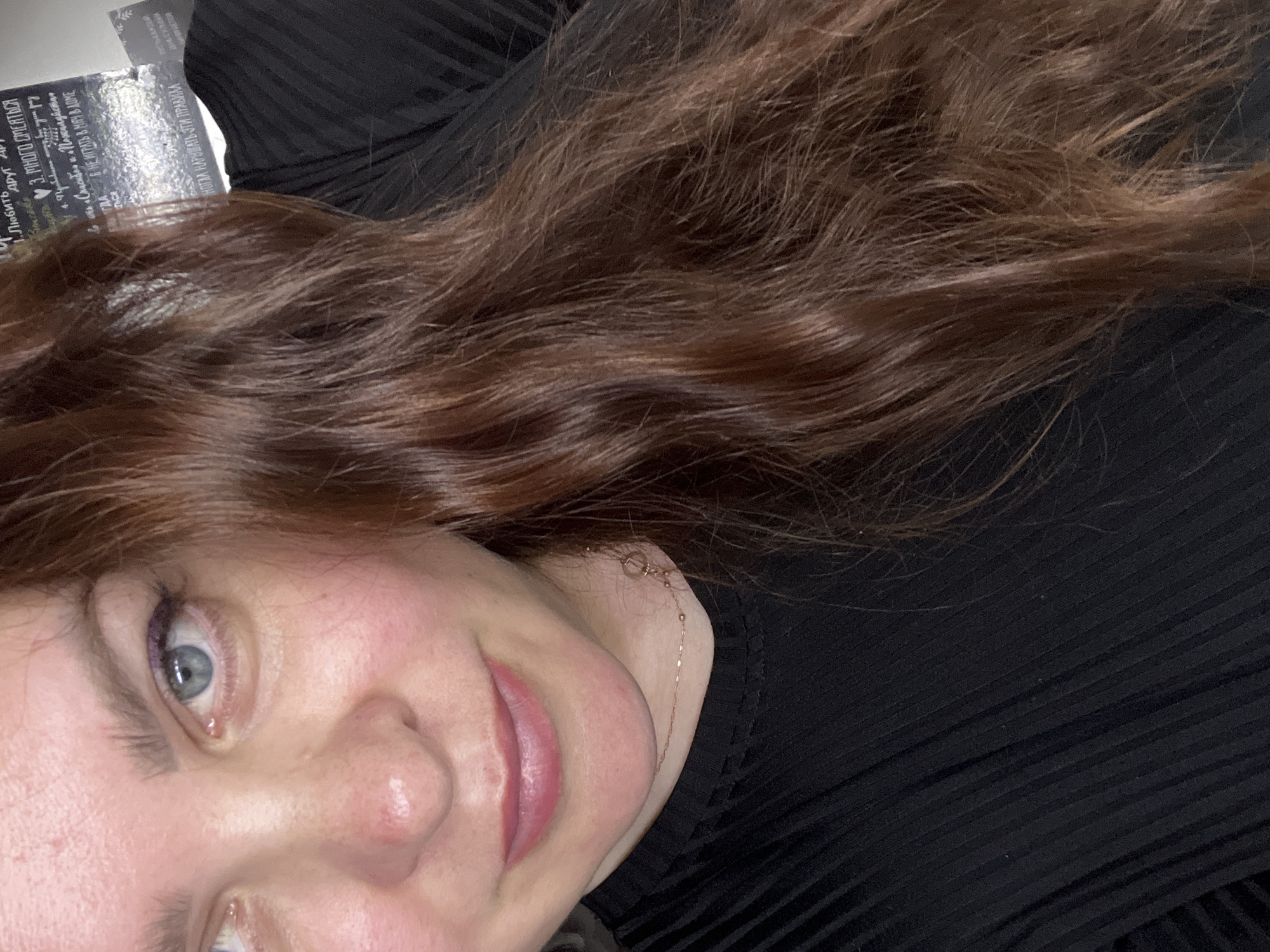 A close-up, chest-up indoor portrait depicts a light-skinned female subject, approximately half of her face visible, looking directly towards the camera with a subtle smile. Her left eye (viewer's right) is fully visible, featuring a light blue iris, a dark pupil, and a subtle application of purple eyeliner on the upper lash line. Her eyebrows are light brown and well-groomed. The skin on her visible cheek and nose shows a reddish undertone. Her lips are full, pink, and appear slightly glossy. Her chin is visible.
Her hair is medium to dark brown with reddish undertones, long, and displays a prominent wavy texture. It cascades down the right side of the frame, partially obscuring her face and extending across her chest.
The subject is wearing a black, finely ribbed, long-sleeved top. A delicate, rose-gold toned chain necklace with small, irregularly spaced metallic elements and a circular pendant or clasp detail is visible around her neck.
The background, predominantly behind the subject's left side (viewer's right) and partially obscured by her hair, is out of focus. It features a dark surface with handwritten white Cyrillic text. Recognizable phrases include a numbered list, with entries such as "1. ЛЮБИТЬ" (1. LOVE), "3. МНОГО СМЕЯТЬСЯ" (3. LAUGH A LOT), and "6. НЕ ИГРАТЬ В МЯЧ В ДОМЕ" (6. DON'T PLAY BALL IN THE HOUSE). Below the list, the text "НЕЛЬЗЯ НАРУШАТЬ ЭТИ ПРАВИЛА" (CANNOT VIOLATE THESE RULES) is clearly visible.
The scene is an indoor environment, likely a personal residence or casual establishment, located in the city of Noviy, Russia. Lighting appears soft and consistent, indicative of an interior setting.