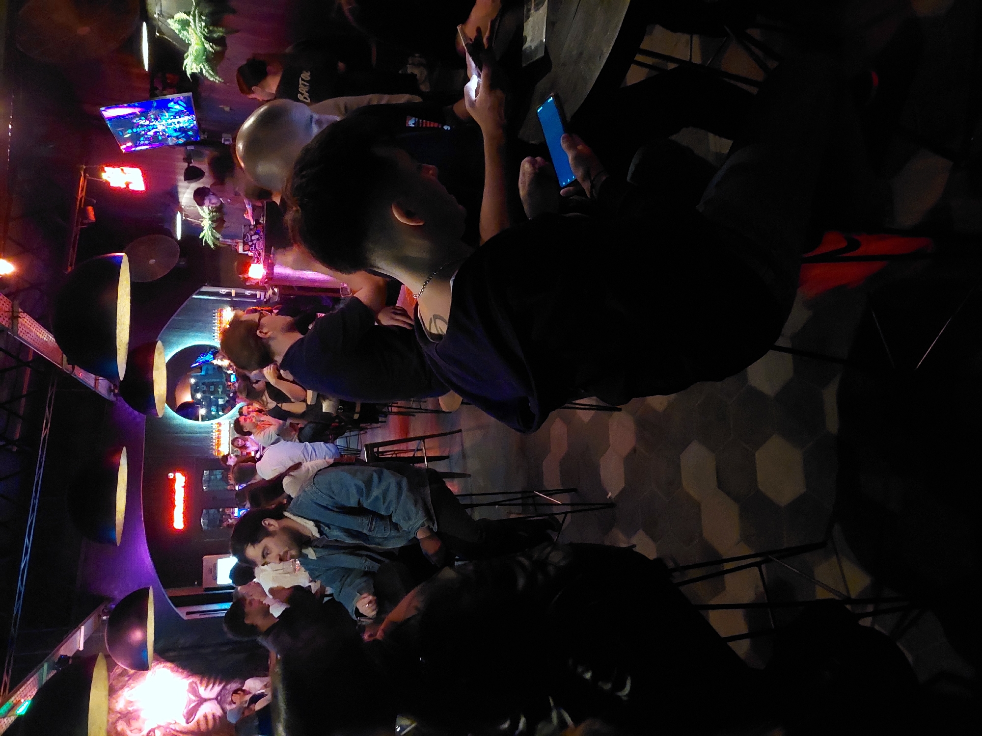 The image shows the interior of a crowded bar, likely in Buenos Aires, Argentina, based on the general ambiance. Numerous patrons are seated on stools at high tables. Some individuals are using their smartphones, while others are engaged in conversation. The lighting is dim and features colorful neon accents, typical of nightlife venues. Overhead, several dome-shaped light fixtures are visible. A television screen displays content in the background. The floor has a hexagonal pattern. The general atmosphere suggests a social gathering in a dimly lit, urban bar setting.