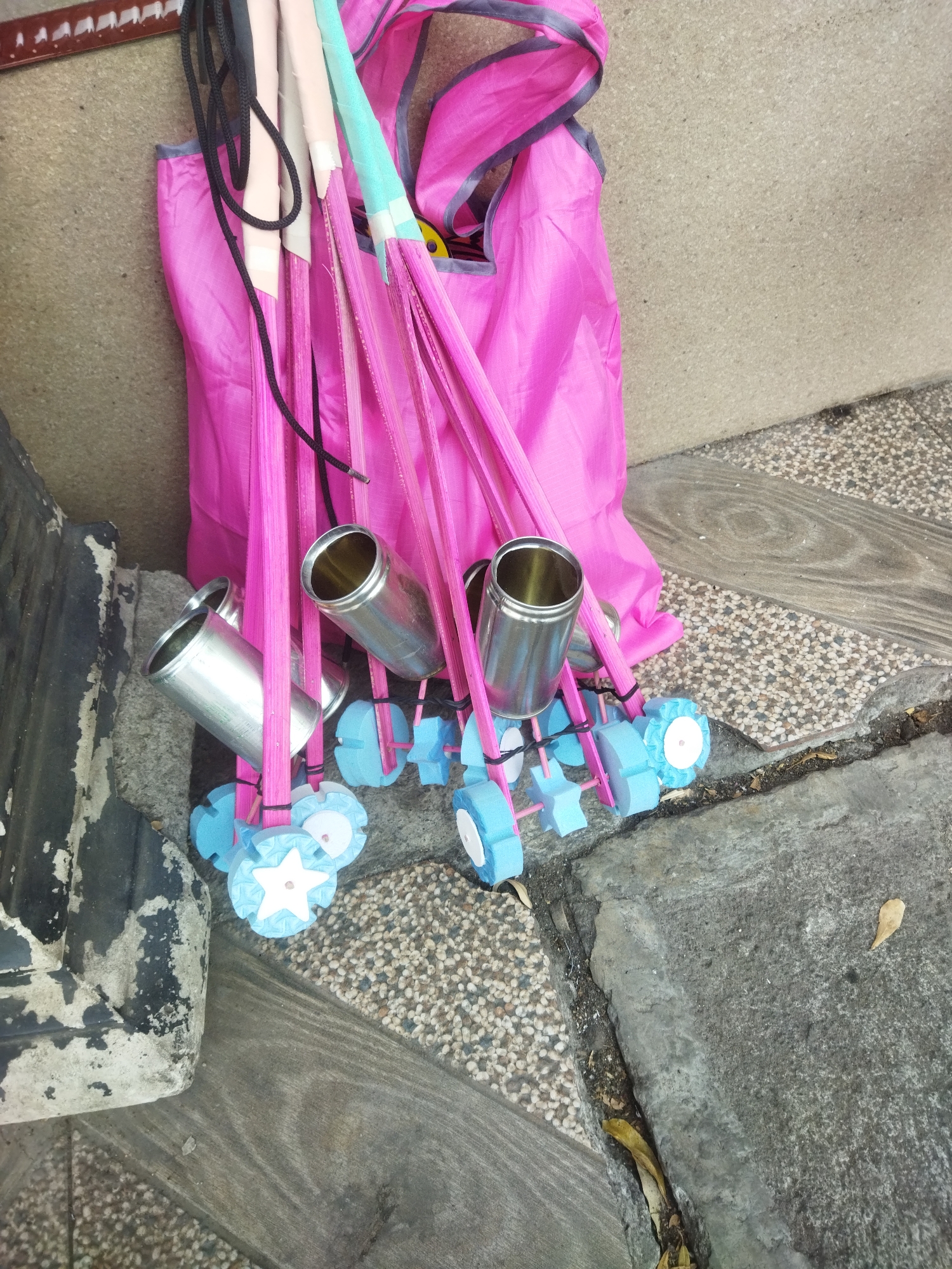 In Wonogiri, Indonesia, a close-up, eye-level shot captures a collection of bright pink and light blue items leaning against a textured concrete wall. A large, vibrant pink bag is the dominant feature, partially obscuring a bundle of thin, pink poles. Several of these poles are capped with metallic silver cylinders, and others are adorned with blue, star-shaped, foam-like attachments, suggesting they might be part of a dismantled play structure or hobby equipment. The ground beneath is a mosaic of small, light-colored pebbles and weathered wooden planks, interspersed with cracks and debris, indicative of an outdoor urban setting. The overall scene is static, with no individuals present.