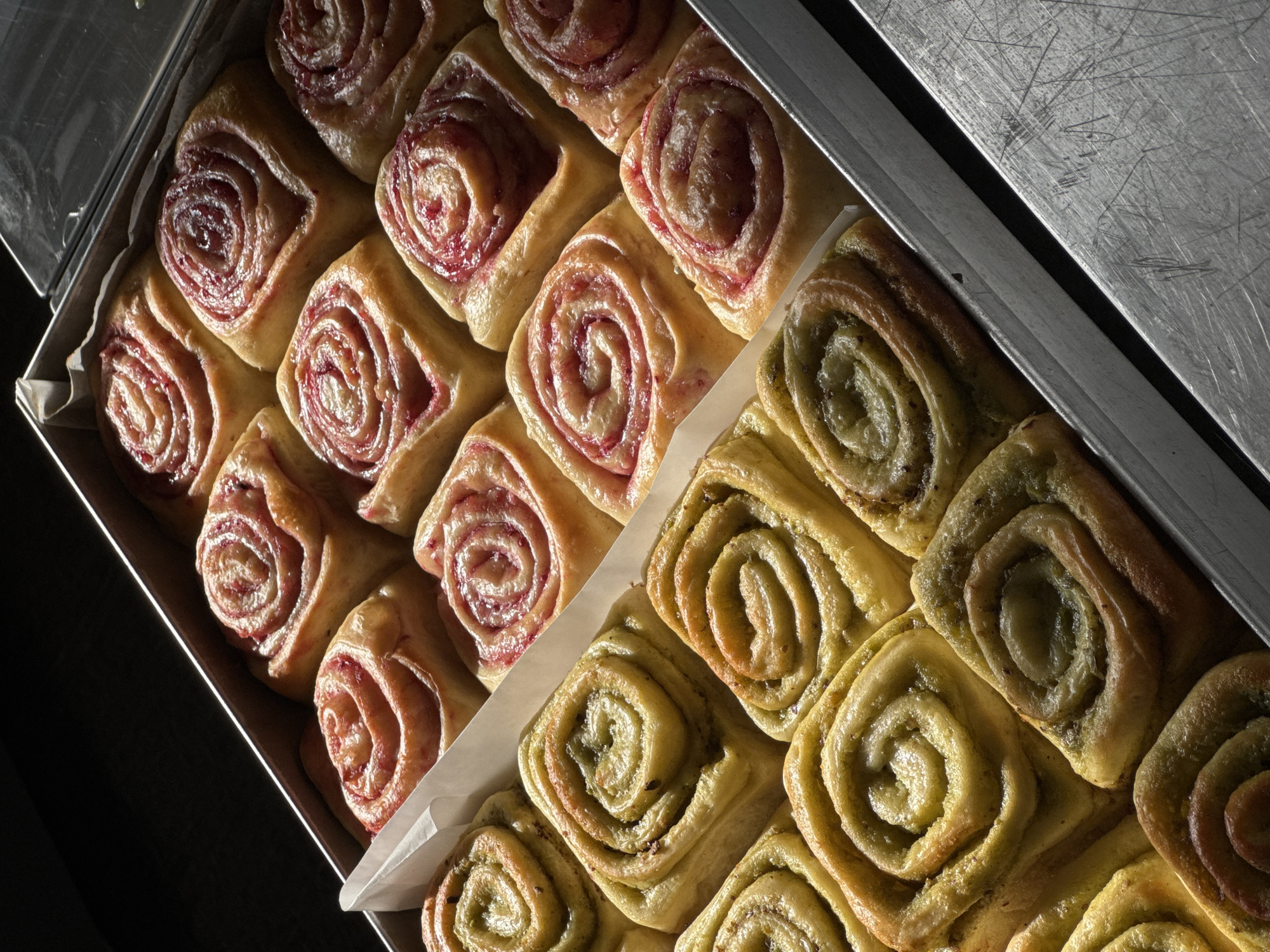 Two rows of freshly baked sweet rolls are arranged in a metal baking pan. The front row consists of green-tinted rolls, likely flavored with matcha or pistachio. The back row features pinkish-red rolls, suggesting a berry or fruit filling. The rolls are golden brown and appear to have a slightly glossy glaze. The pan is positioned on a scratched metal surface, possibly a kitchen counter or workstation. The lighting is dramatic, with strong shadows suggesting a single light source from the upper left. The background is dark and out of focus. The scene appears to be indoors, likely in a bakery or home kitchen. There are no people visible in the image. The origin of this image is not Odesa, Ukraine.