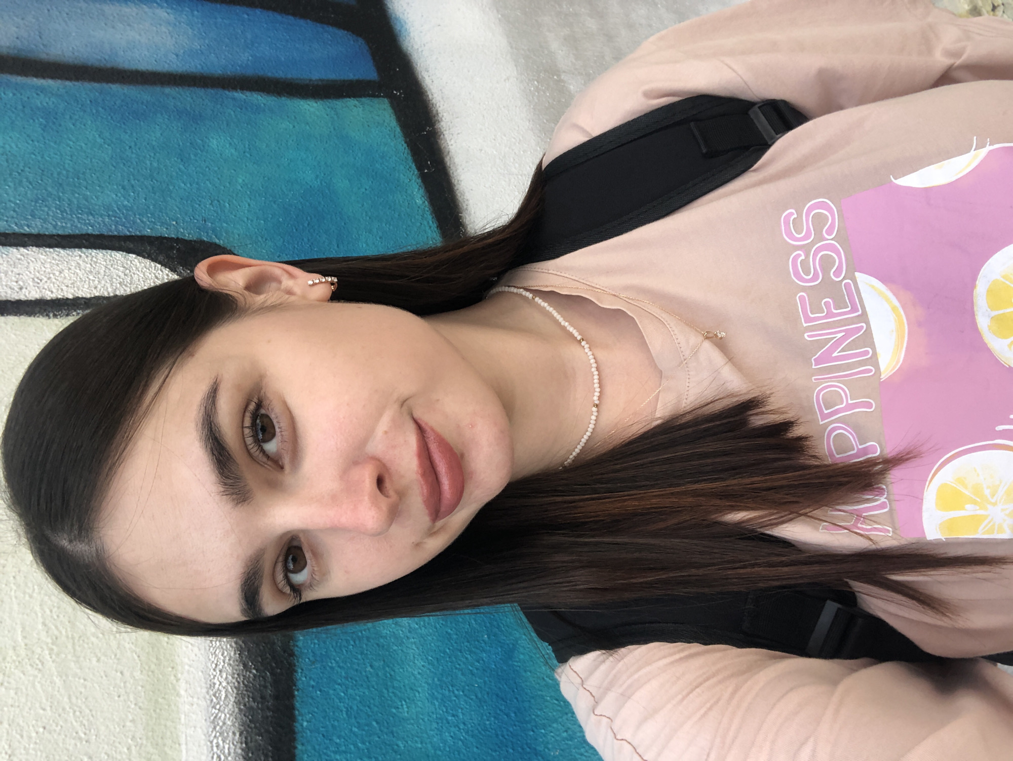 This is a close-up, portrait-style selfie of a young woman with long, dark brown hair, wearing a light pink t-shirt with a graphic design. She has a small pearl necklace and an earring with small stones on her left ear. Her hair is mostly covering her left side, and her head is tilted. The background features a vibrant mural with large sections of blue and white, suggesting an outdoor setting. The lighting appears to be natural, and the overall mood is casual and relaxed. The text "HAPPINESS" is visible in pink lettering on her t-shirt. The image is oriented sideways, with the subjects' heads appearing at the bottom. The context provided indicates the location is Chaikovsky, Russia, though this is not visually apparent from the image itself.